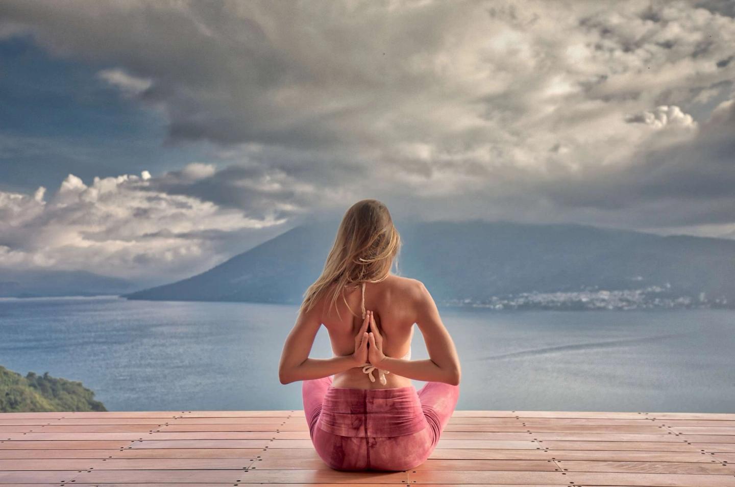 Woman seated in a reverse prayer pose on the Eagle's Nest yoga platform, looking out over Lake Atitlan and a volcano under storm clouds