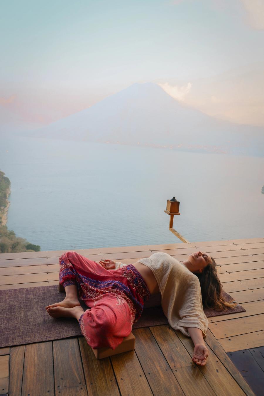 Woman resting in a supported restorative yoga pose on the Eagle's Nest yoga platform, with a hazy volcano over Lake Atitlan behind her