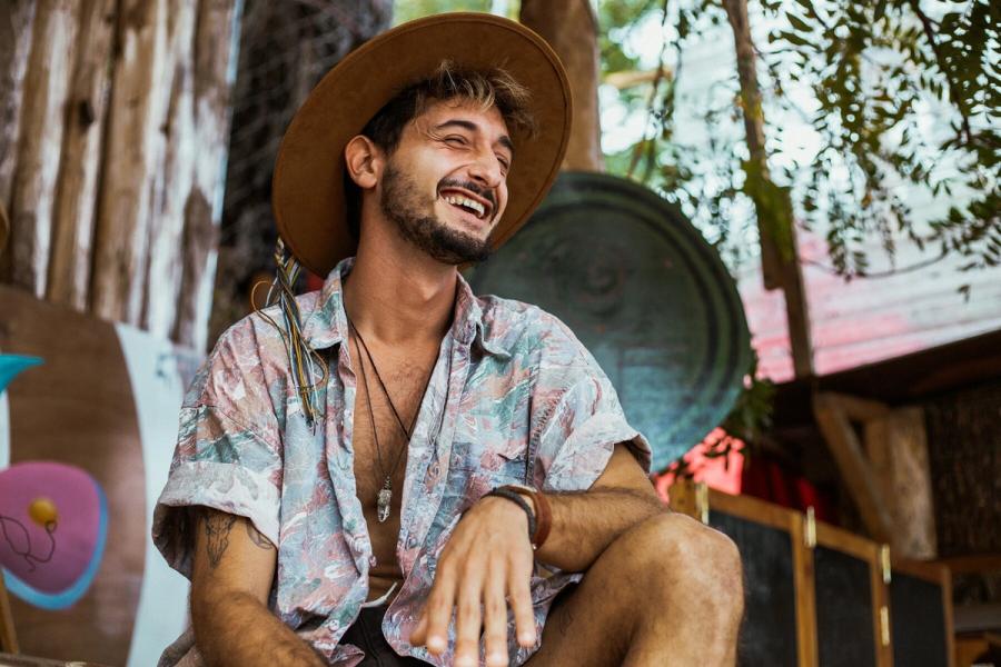 Smiling guest in a brown felt hat and patterned shirt sitting outdoors at Eagle's Nest, with a large gong behind him