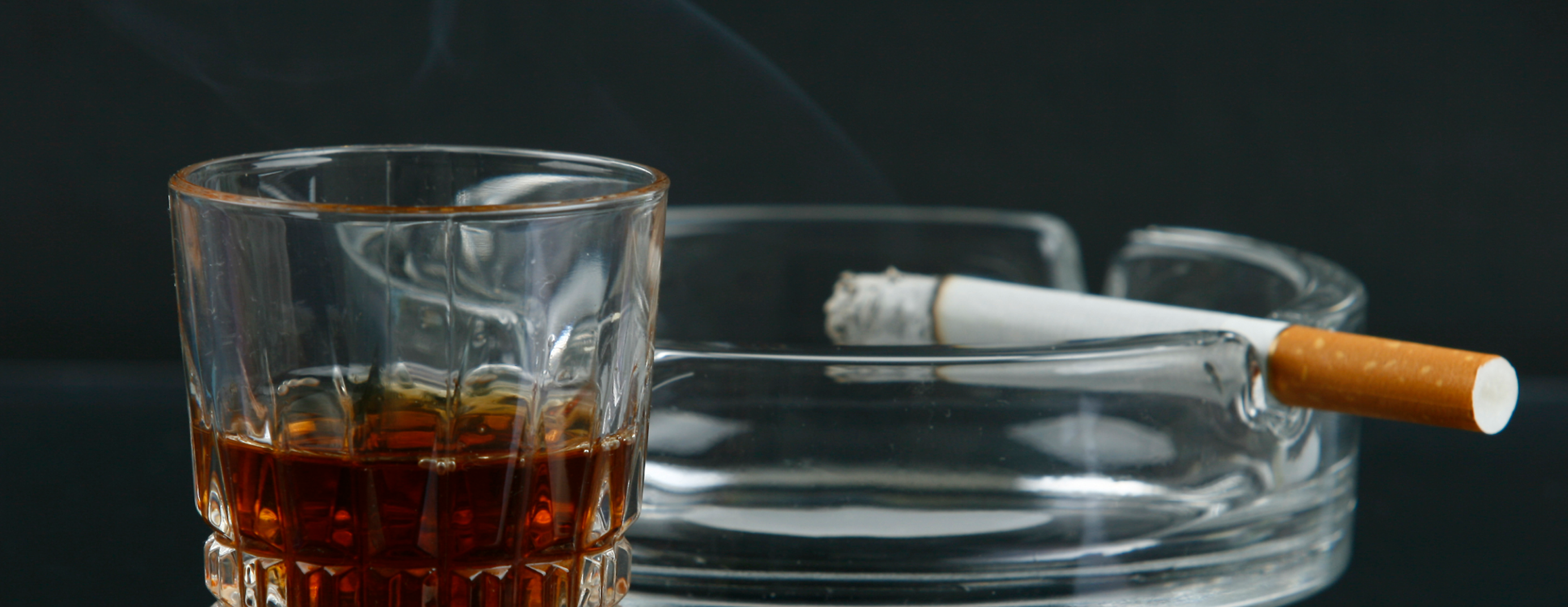 A cup of whiskey next to a cigar laying on an ashtray