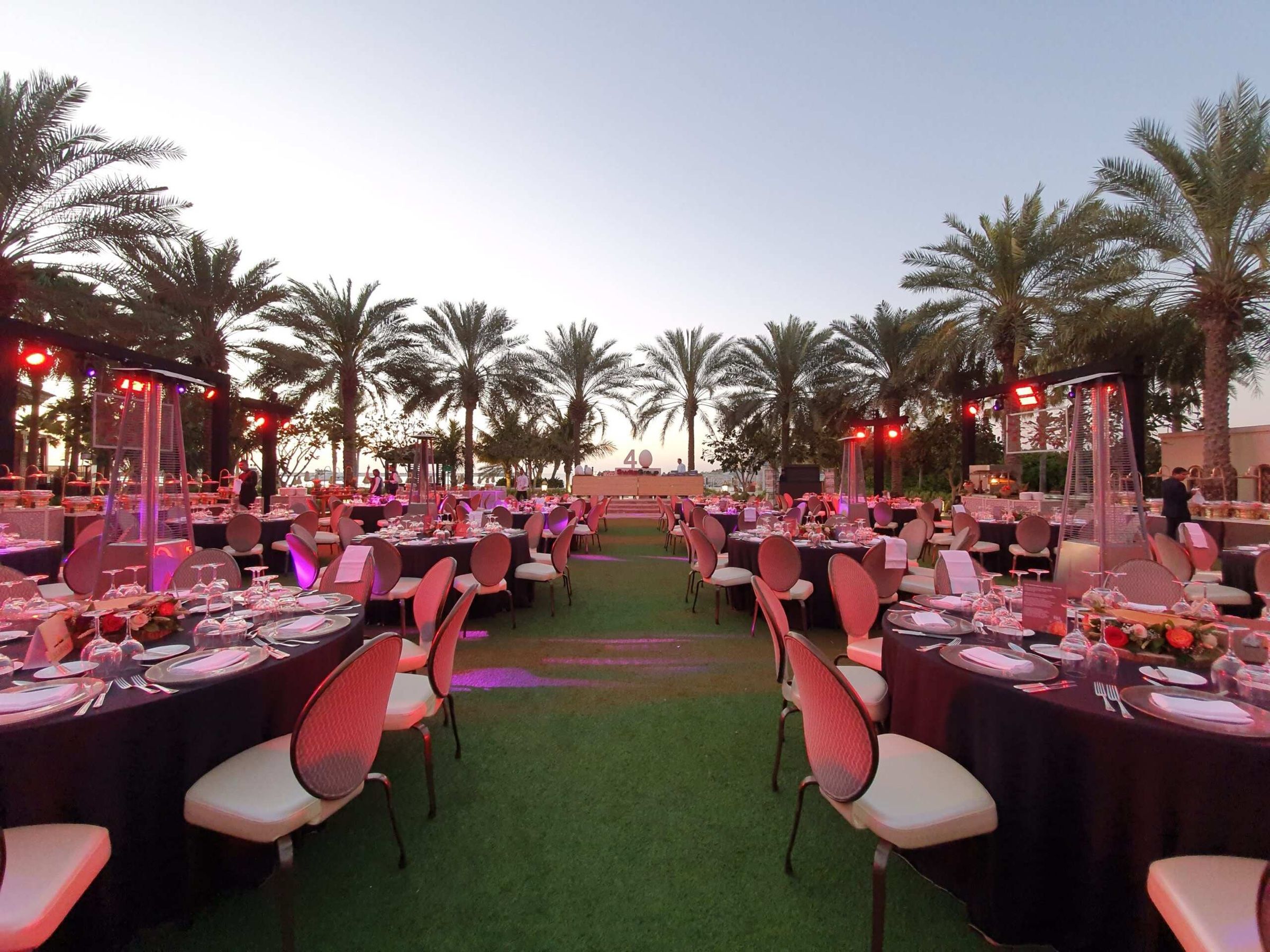Outdoor gala dinner with long tables and pink chairs under palm trees at dusk