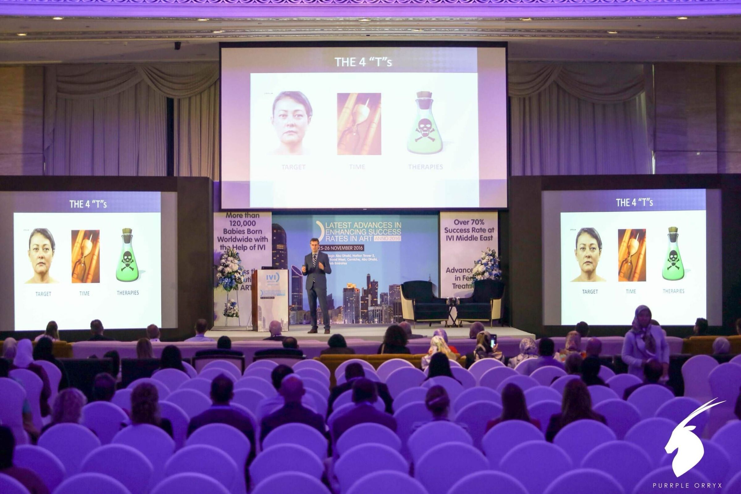 Speaker presenting on stage with three screens and purple audience seating