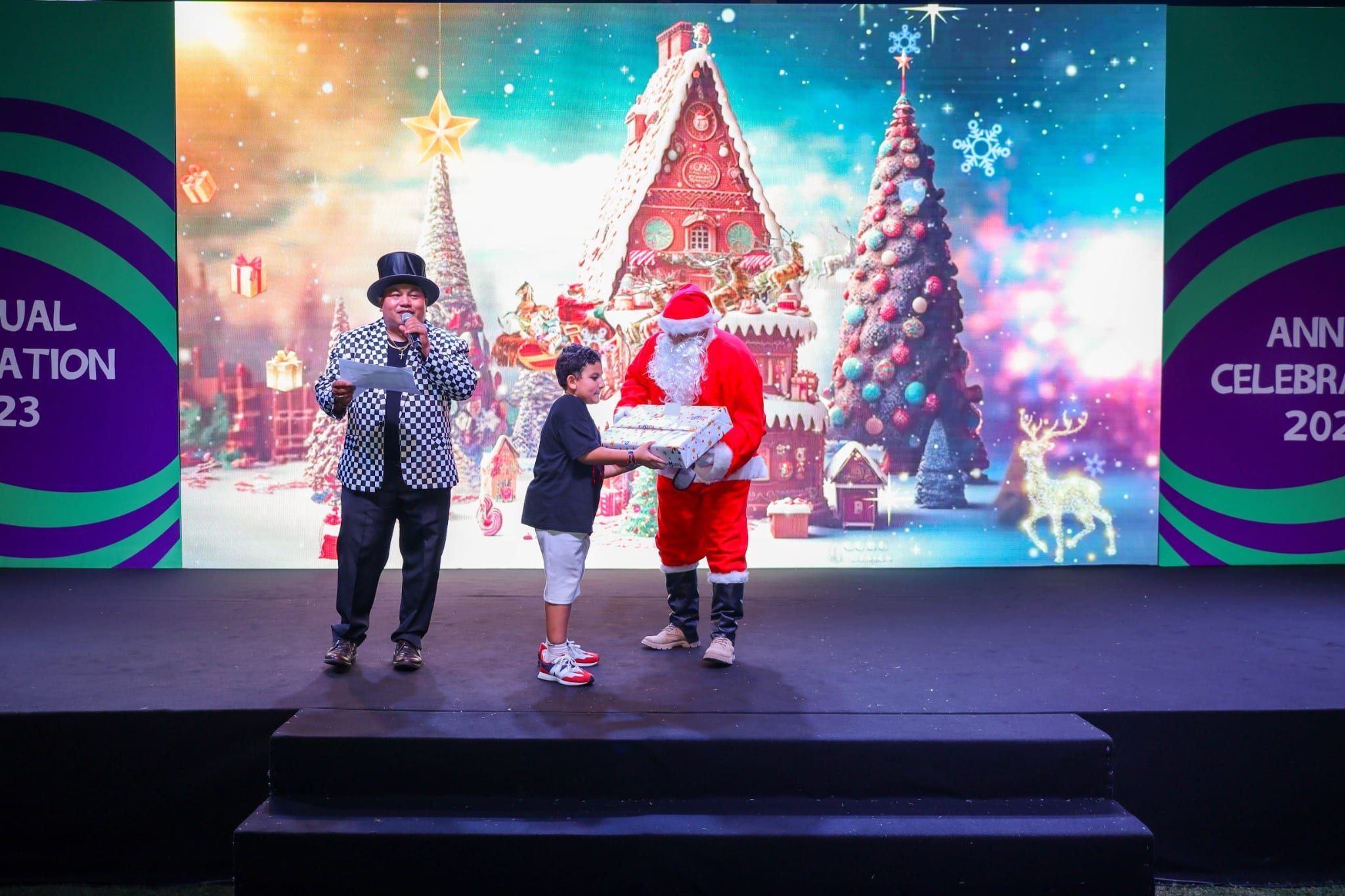 Host and Santa with a child on stage in front of a Christmas village LED backdrop