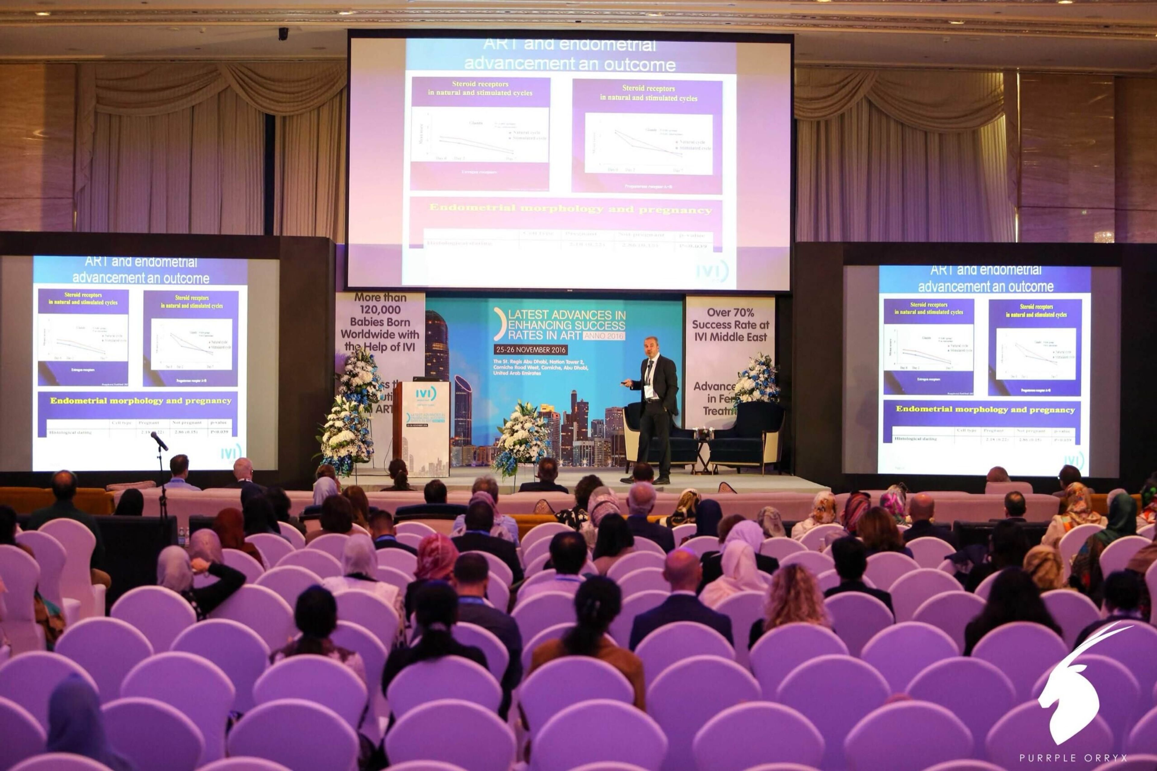 Conference with rows of purple chairs and three presentation screens