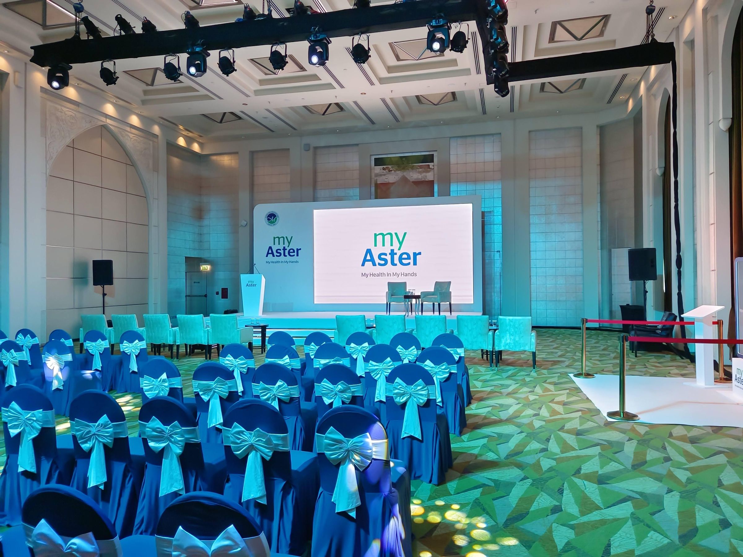 Conference hall with blue-covered chairs facing a branded stage screen