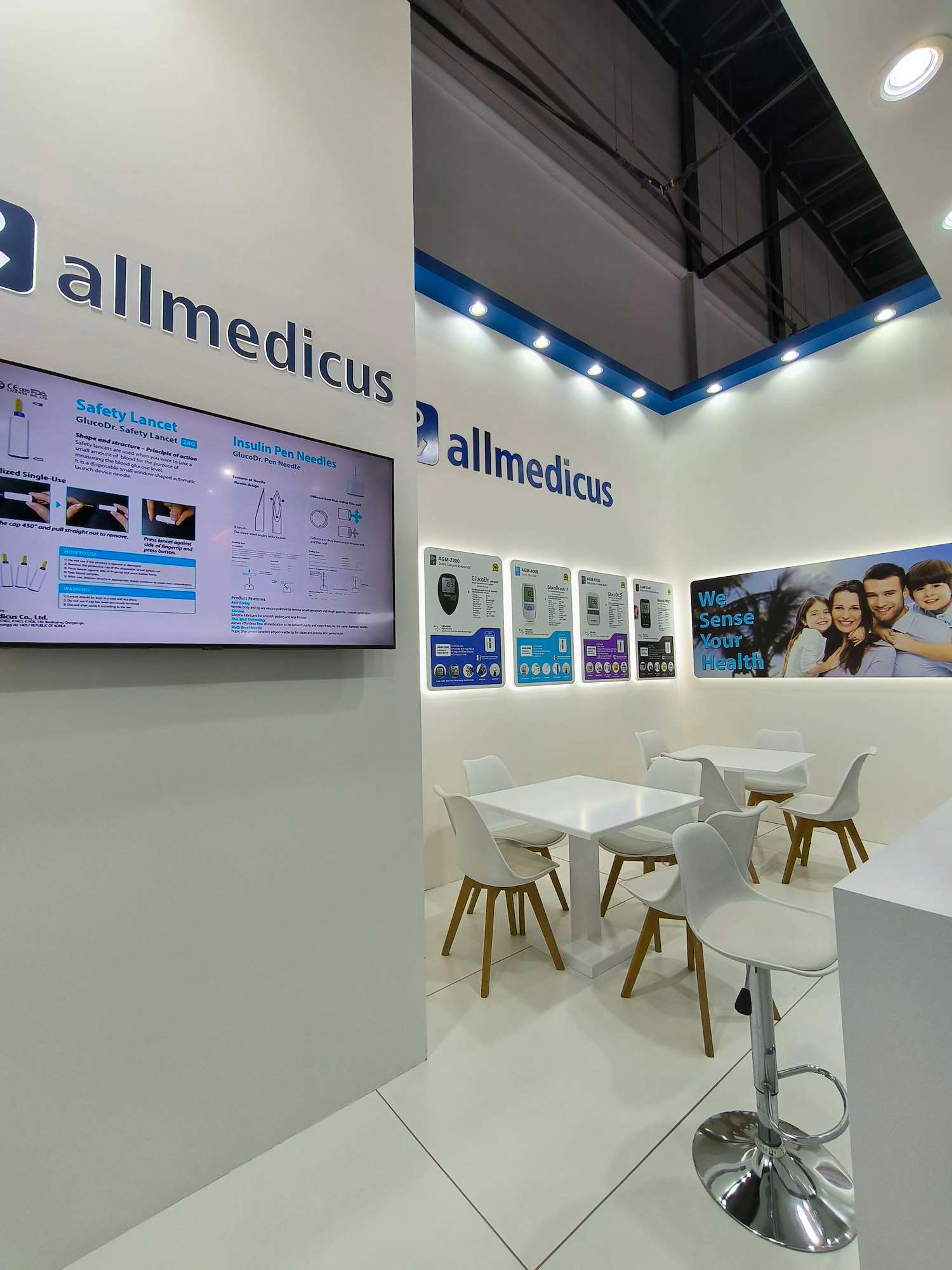 White exhibition booth with blue-edged product panels, a small screen, and white tables and chairs at a trade show.