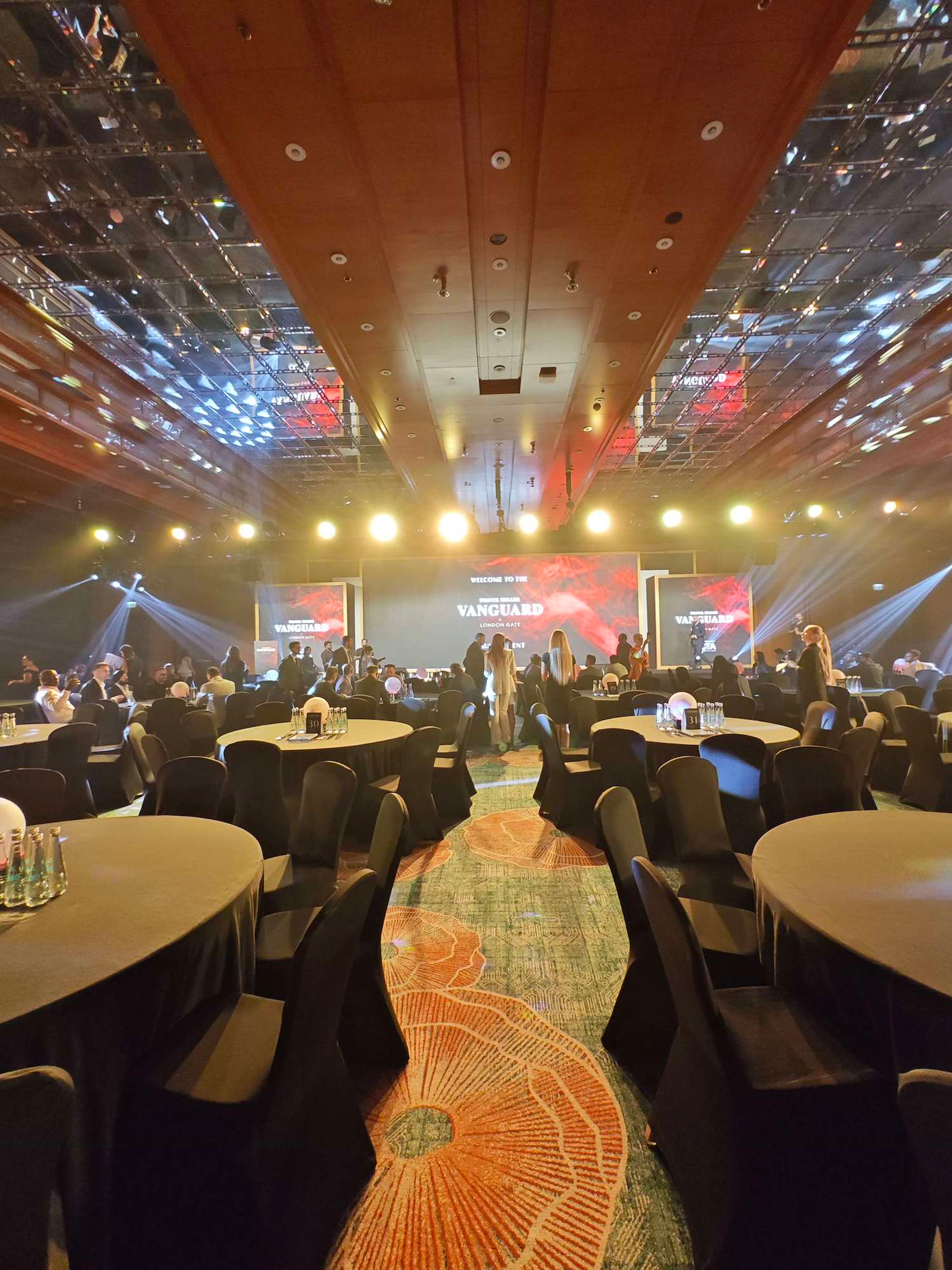 Guests gathering between tables in a ballroom before a product launch, with three LED screens and light beams on stage