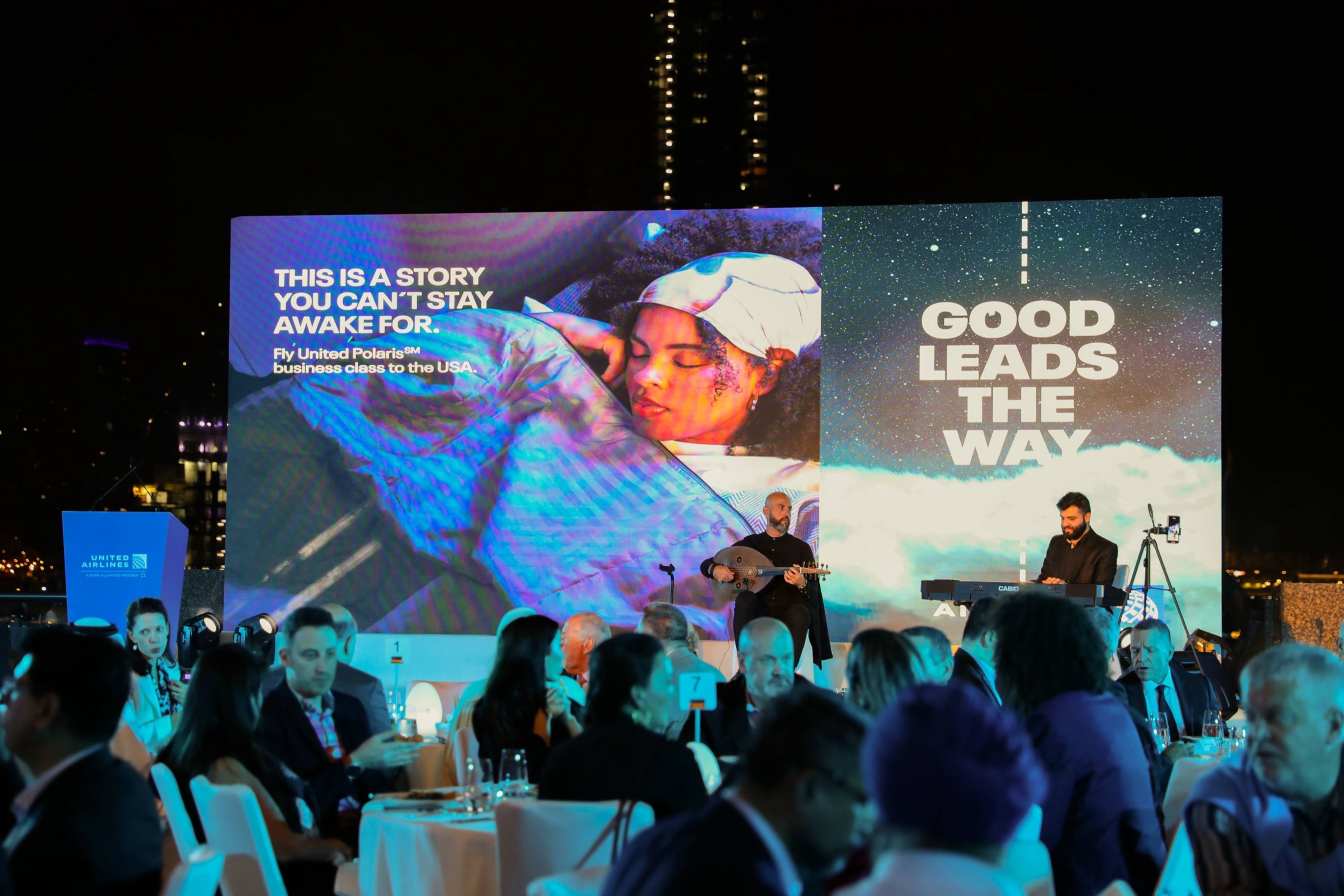 Audience seated in a corporate event, listening to a speaker on stage with large presentation screens displaying the event name and details