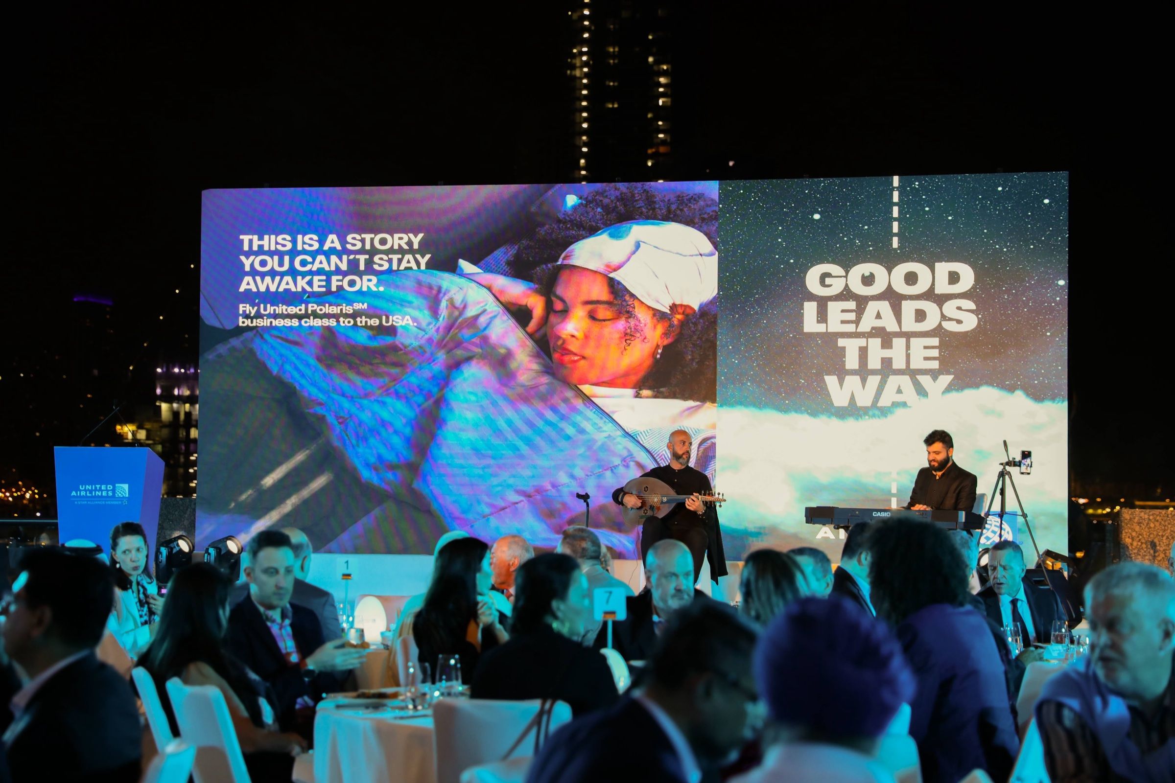 Audience seated in a corporate event, listening to a speaker on stage with large presentation screens displaying the event name and details