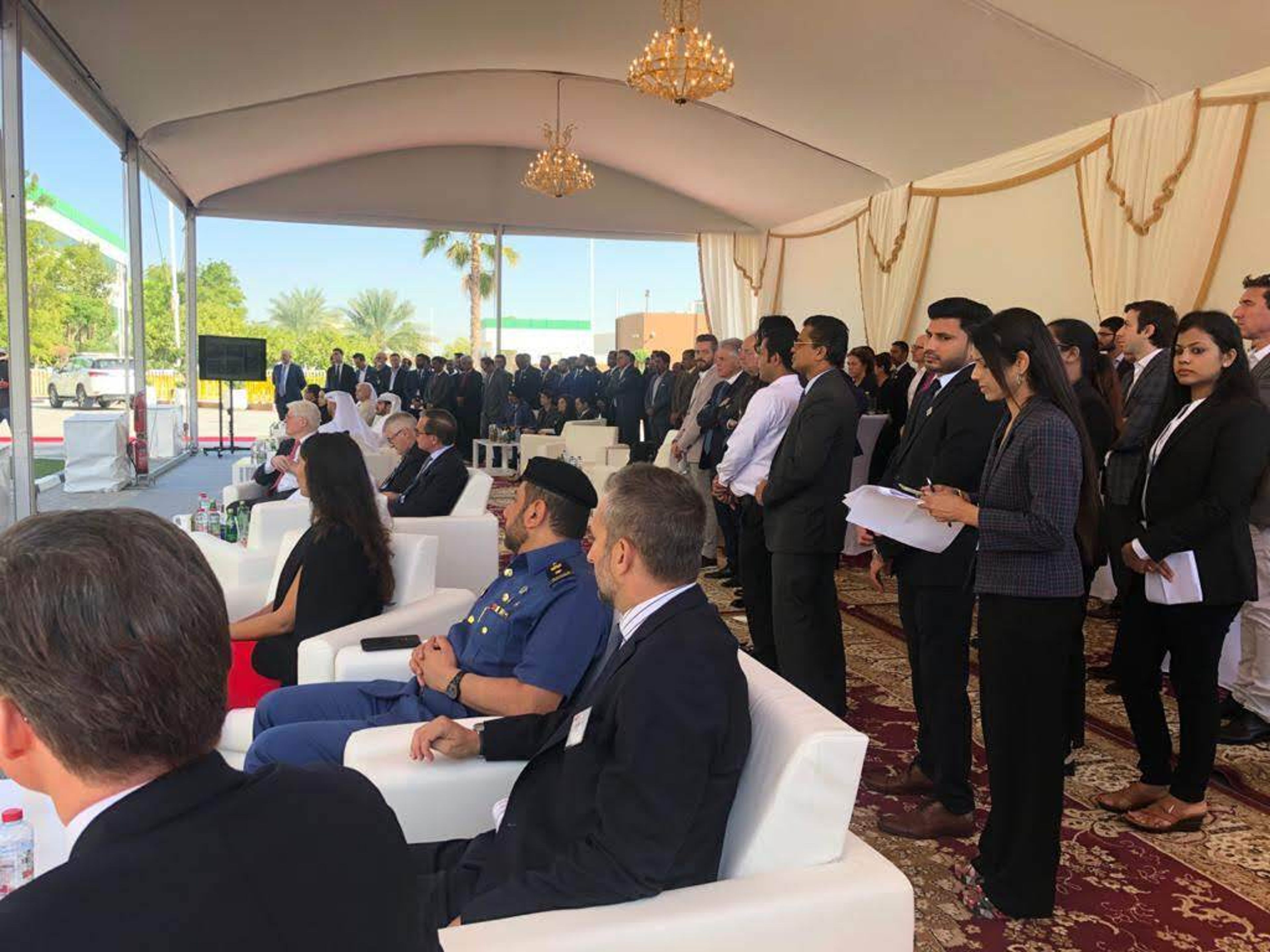 Guests, officials and a uniformed officer seated in a chandelier-lit tent during a corporate opening ceremony