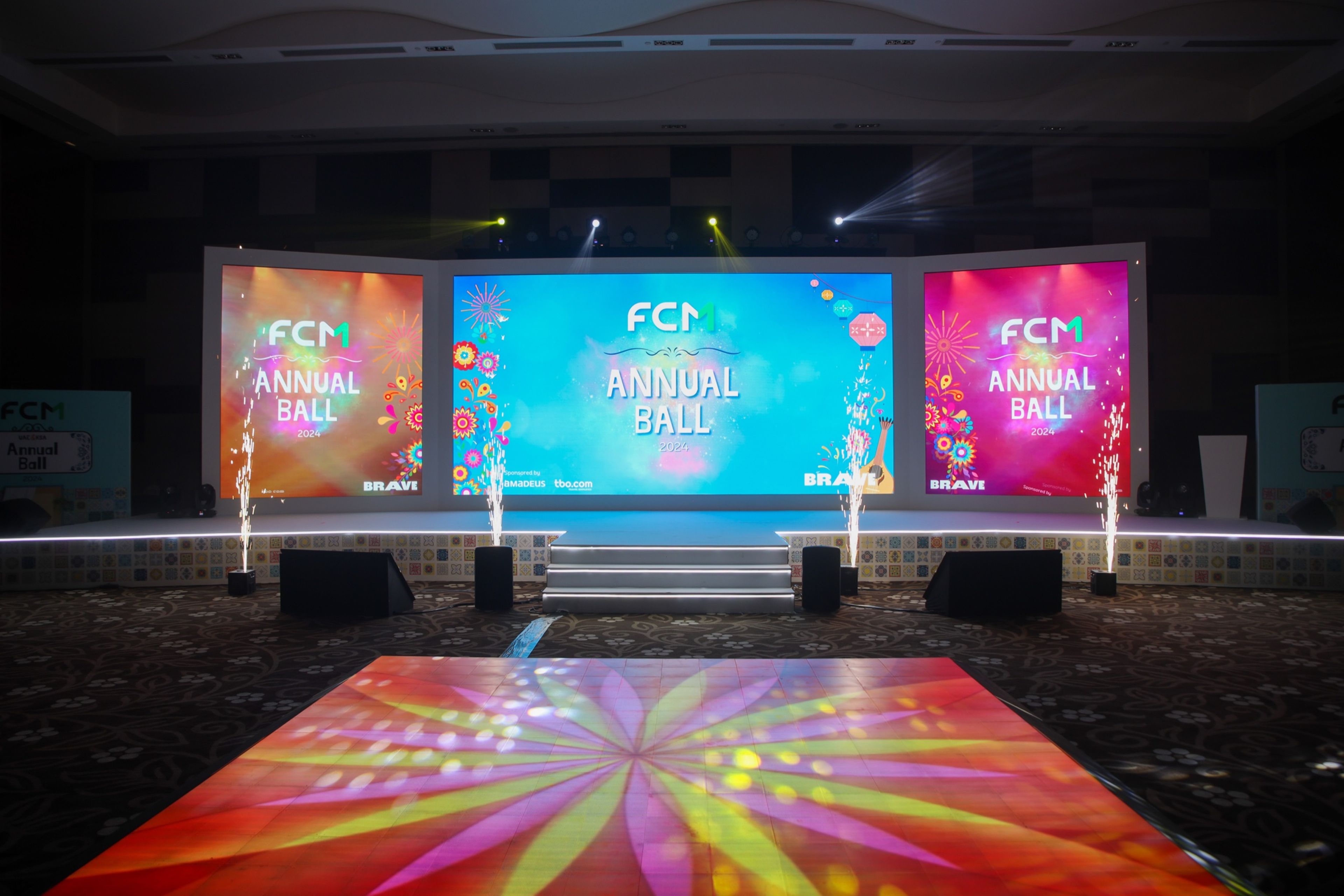 Stage setup for the FCM Annual Ball 2024, featuring colorful digital screens displaying the event name, surrounded by decorative lights and fireworks effects.