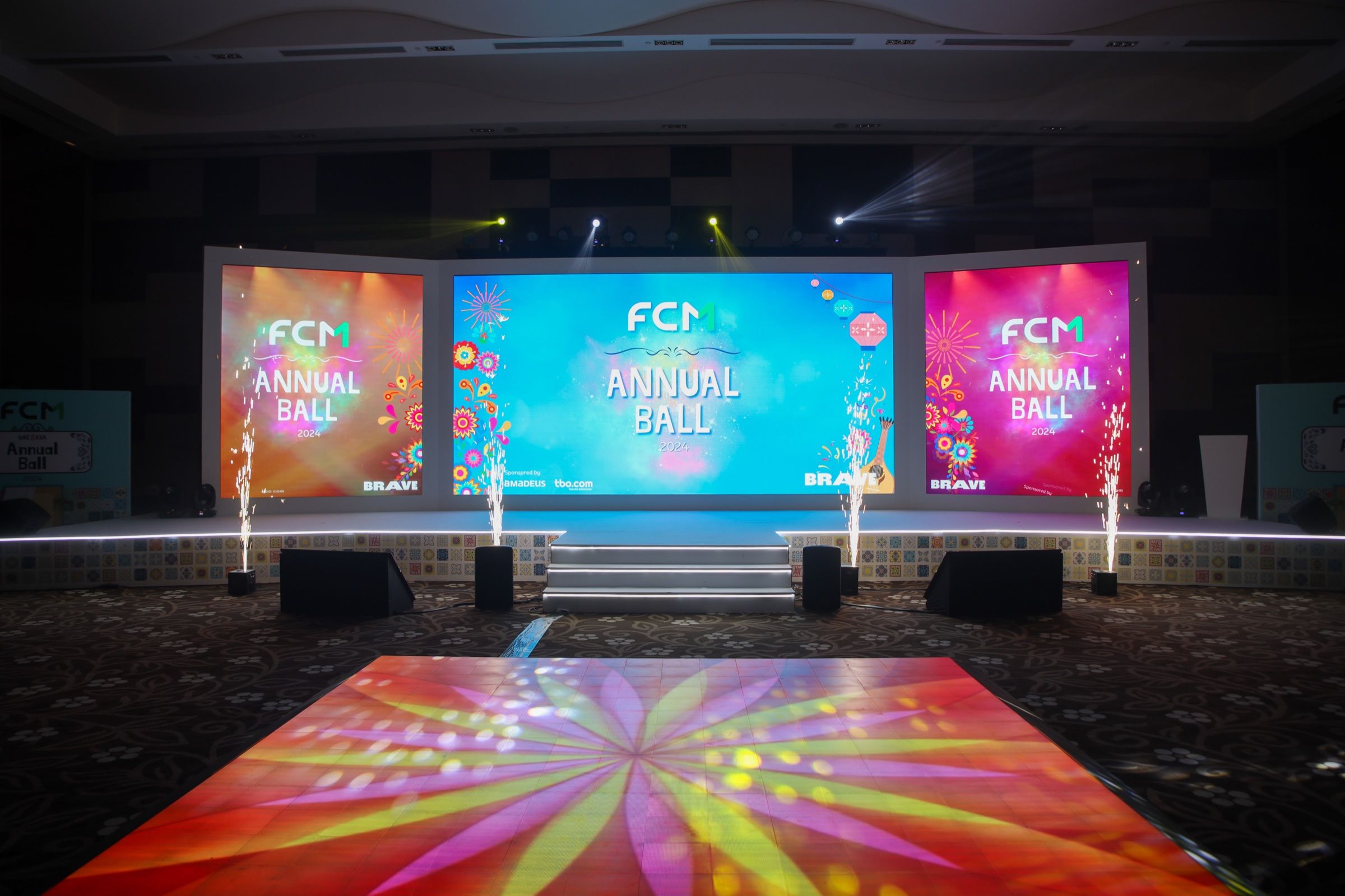 Stage setup for the FCM Annual Ball 2024, featuring colorful digital screens displaying the event name, surrounded by decorative lights and fireworks effects.