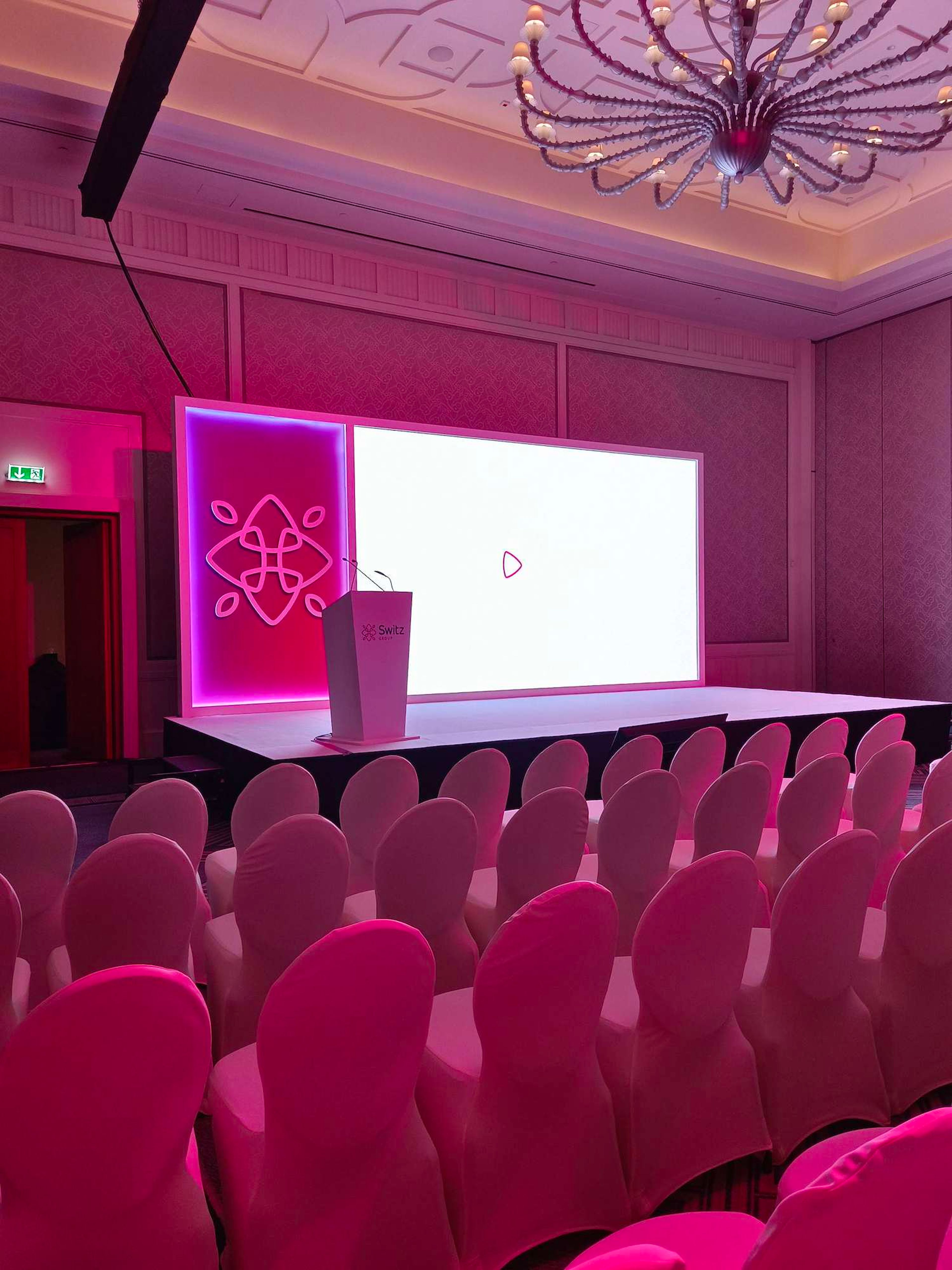 Theatre-style corporate conference setup with LED screen, lit set piece, lectern and covered chairs