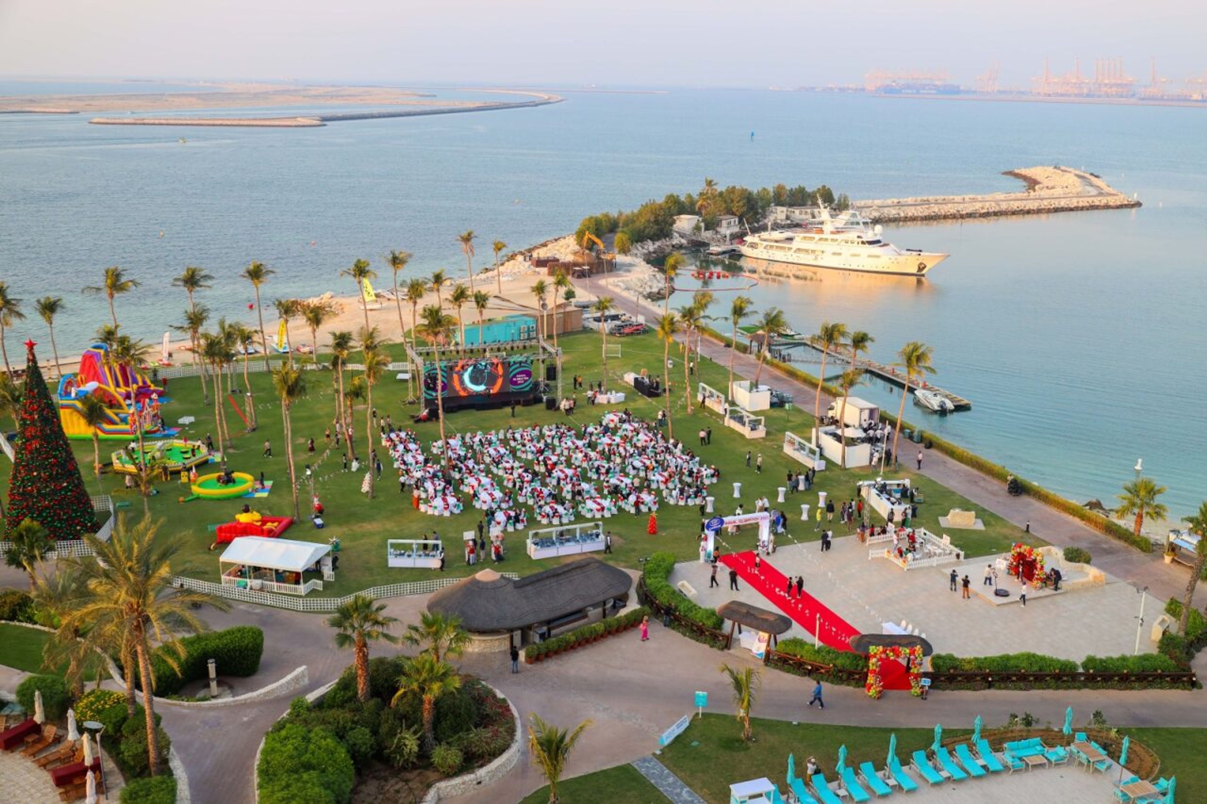 Aerial view of an outdoor Christmas carnival family day on a beachfront resort lawn in Dubai, with a stage, inflatables and guest seating