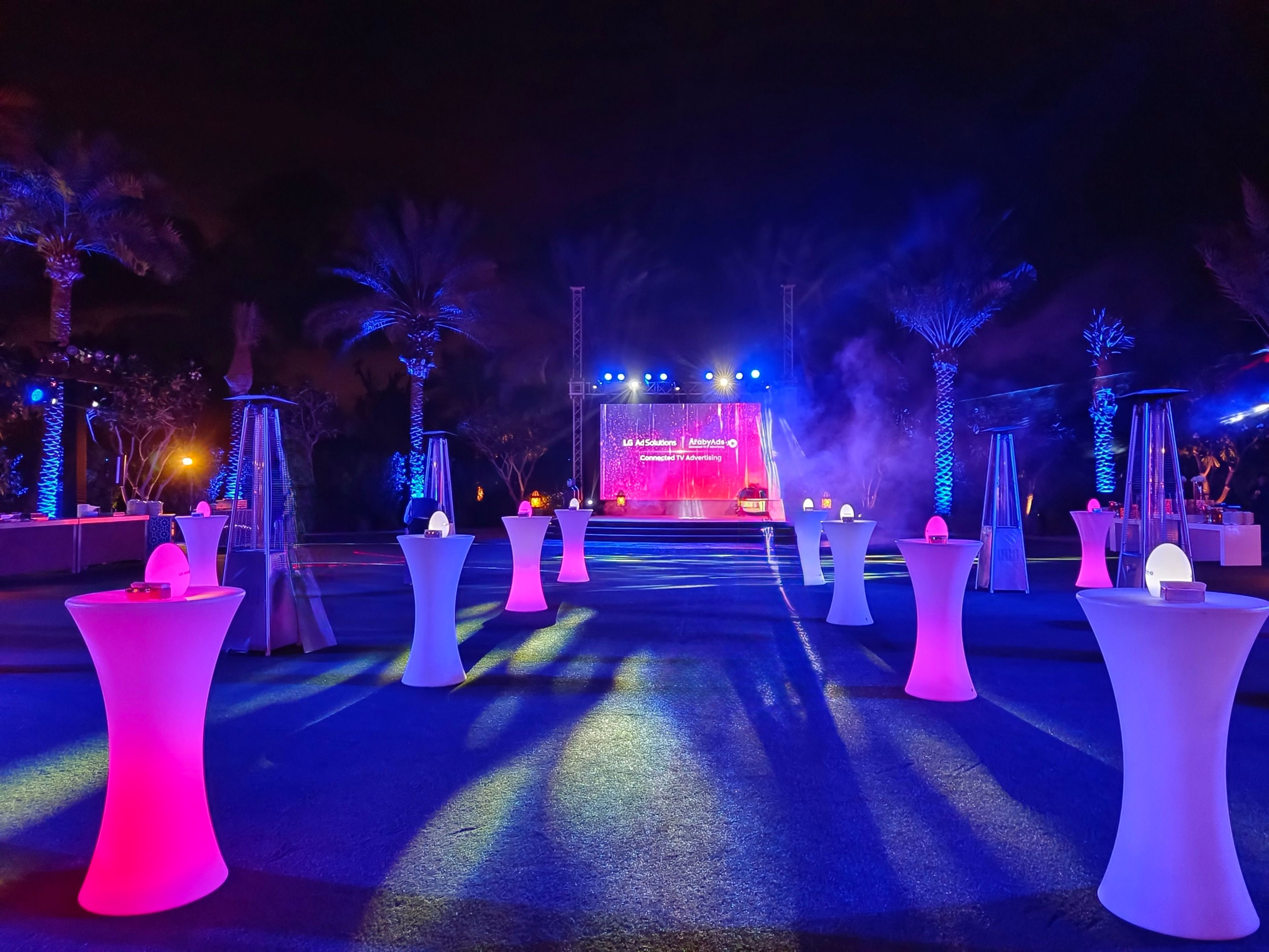 Evening networking event on a lawn with lit cocktail tables, palm trees and a stage screen