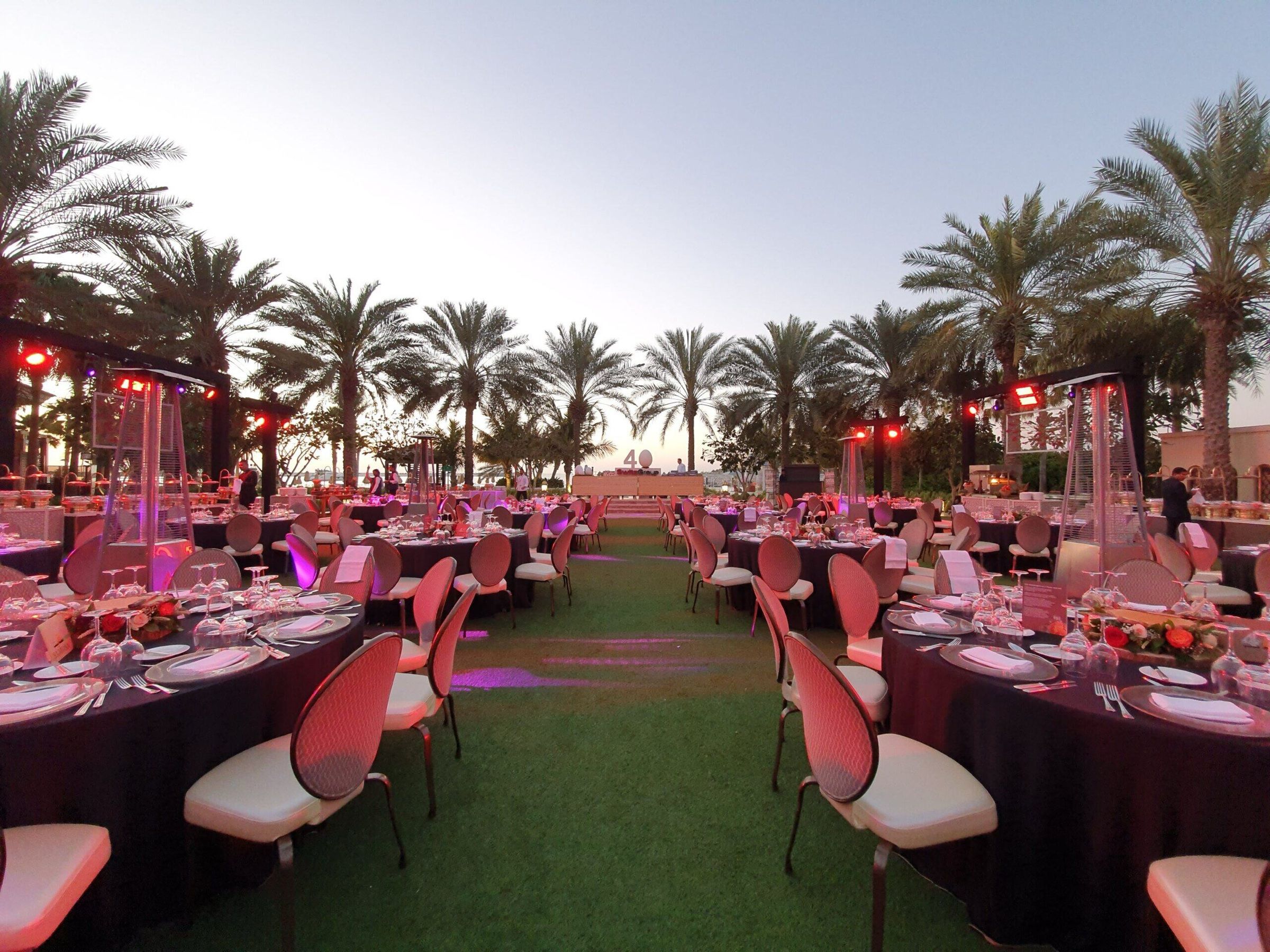 Outdoor gala dinner with long tables and pink chairs under palm trees