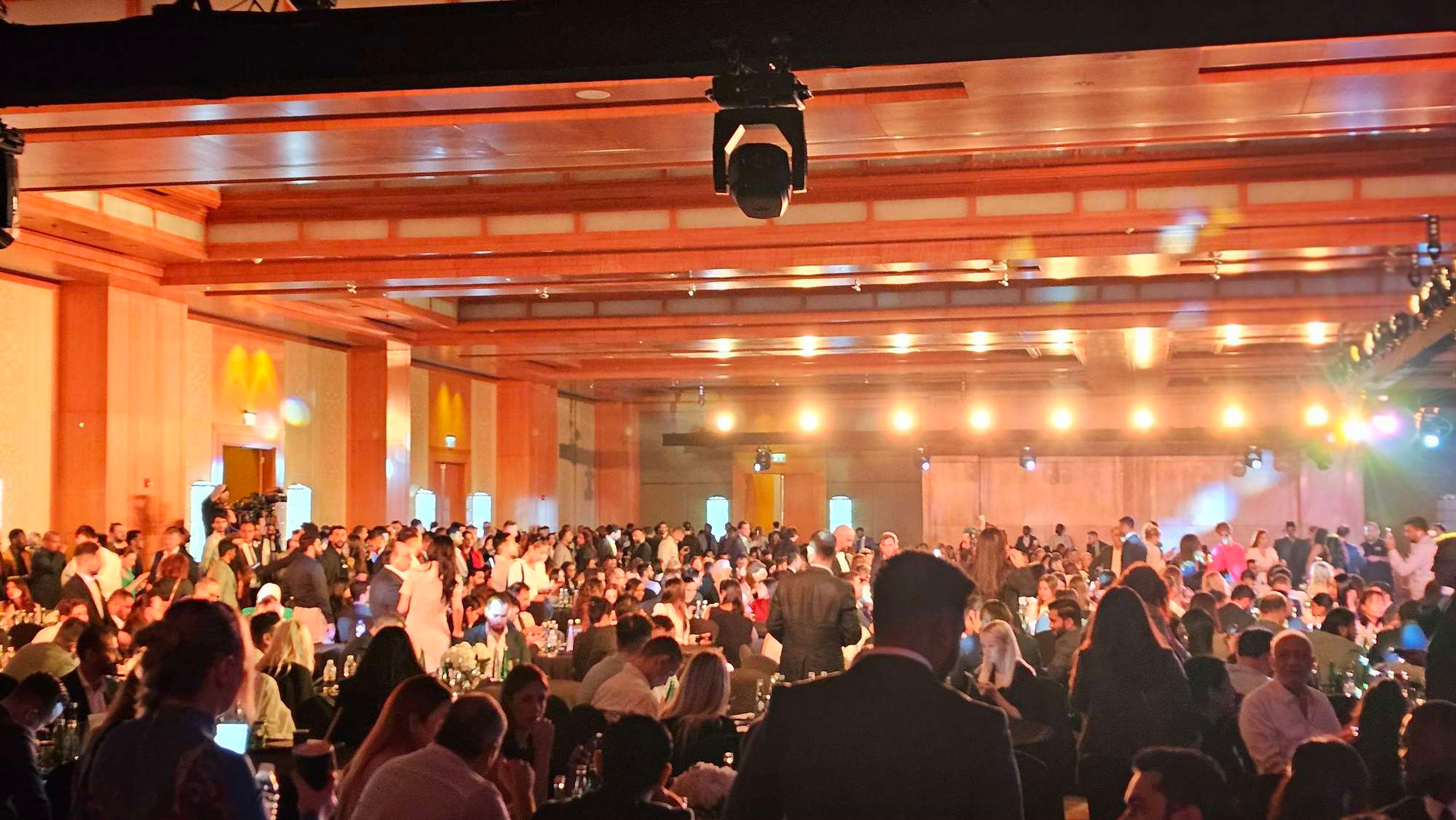 Packed ballroom at a product launch, hundreds of guests seated at tables under rigged lights and a ceiling camera