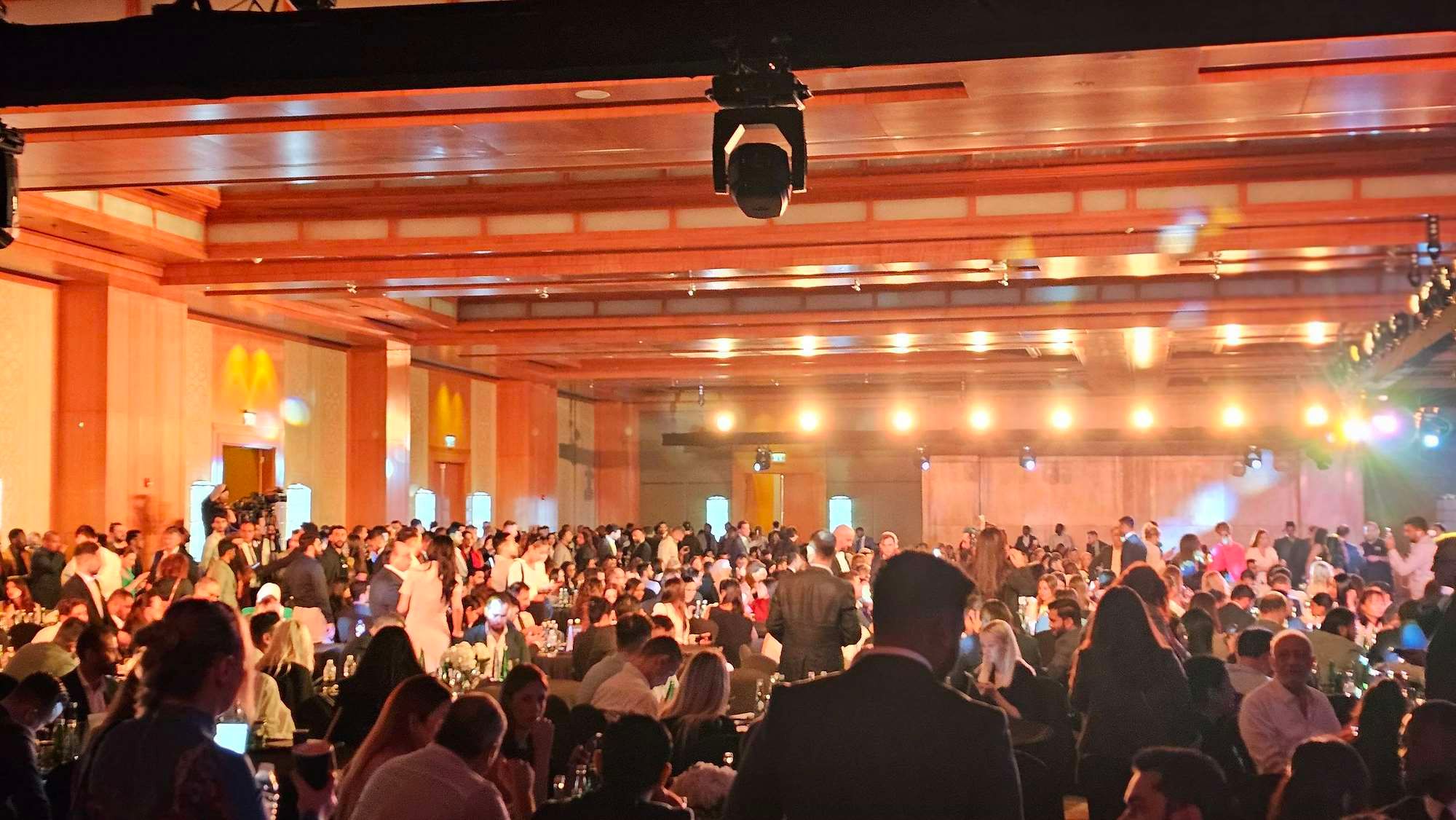 Packed ballroom at a product launch, hundreds of guests seated at tables under rigged lights and a ceiling camera