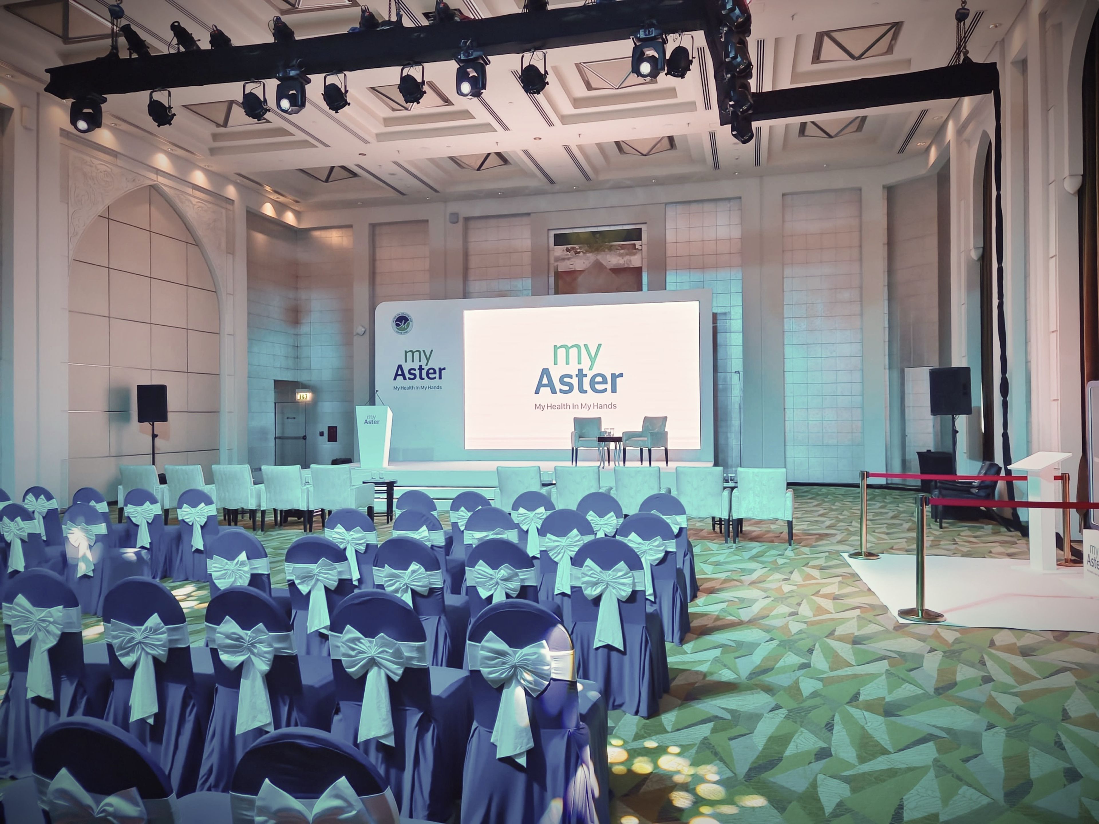 Conference hall with blue-covered chairs facing a branded stage screen