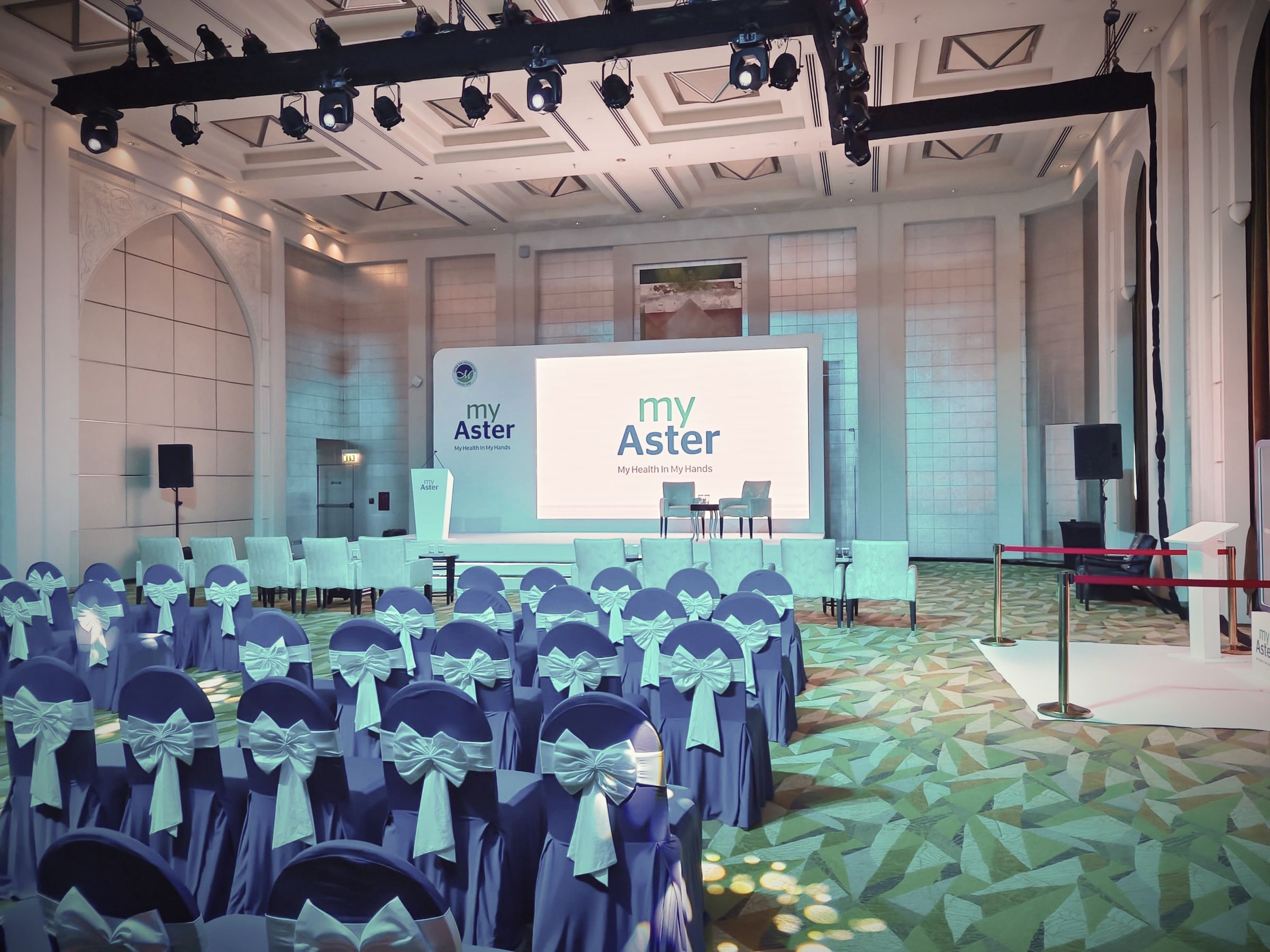 Conference hall with blue-covered chairs facing a branded stage screen