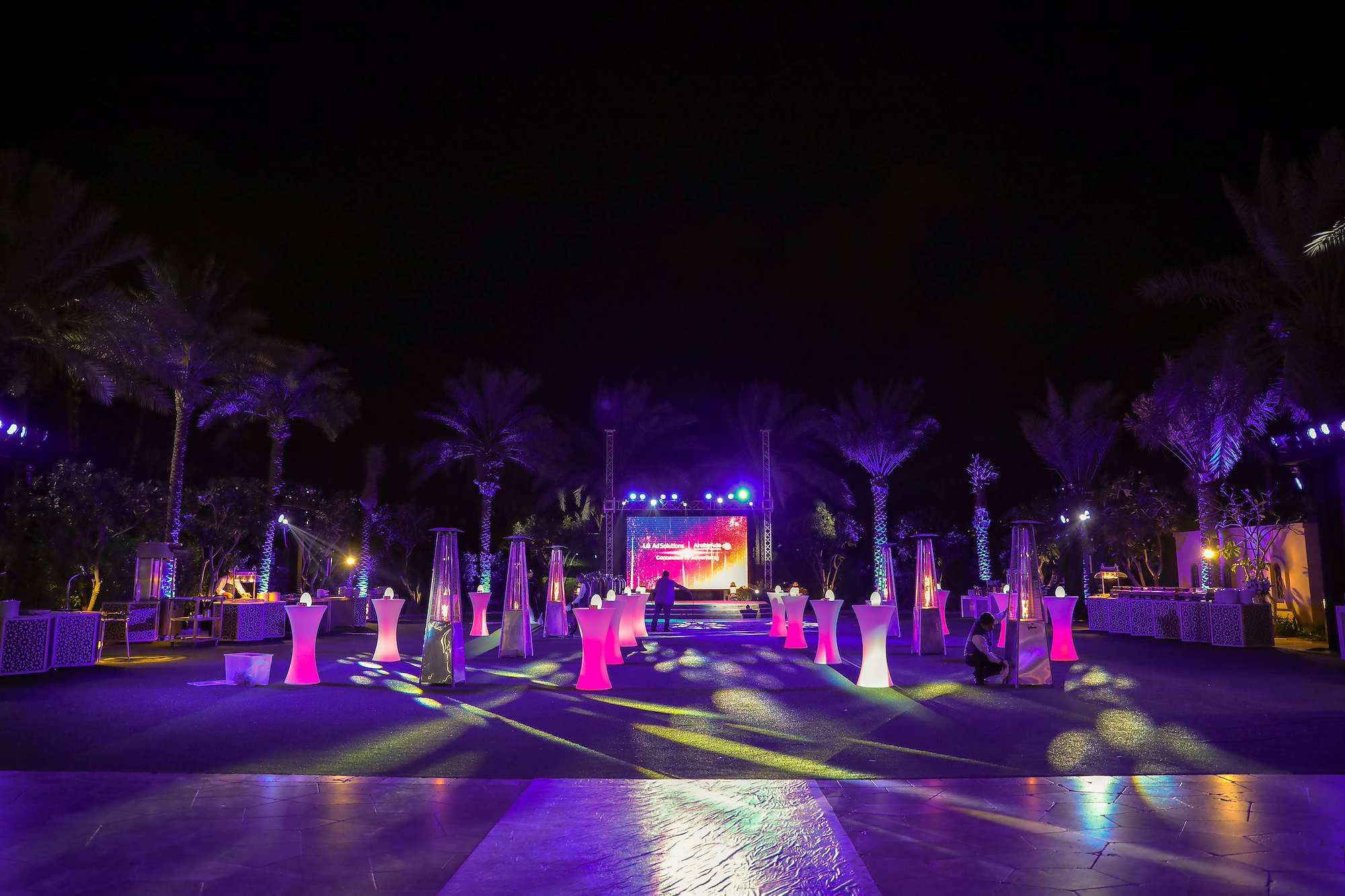 Outdoor corporate event in the UAE with an LED screen on truss, uplit palm trees and lit cocktail tables at night