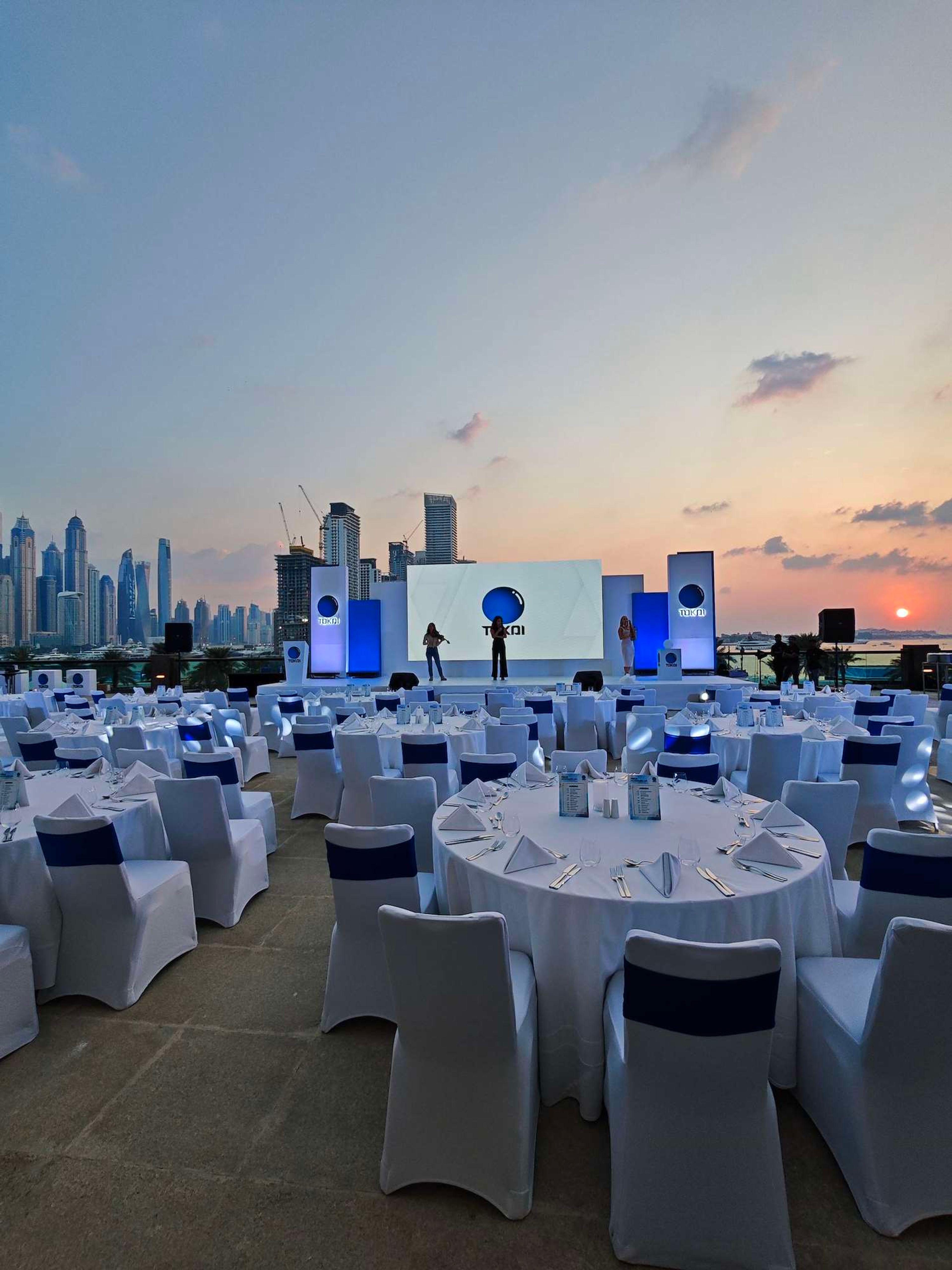 Outdoor rooftop product launch dinner at sunset, white dressed tables facing a stage with a city skyline behind