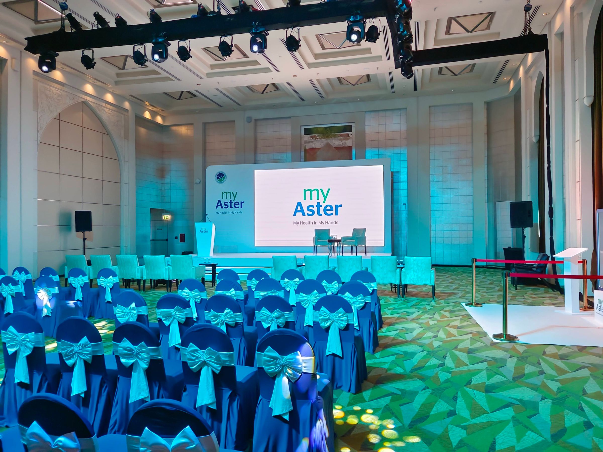 Stage setup for a professional conference, with a large screen displaying event branding and rows of seated attendees