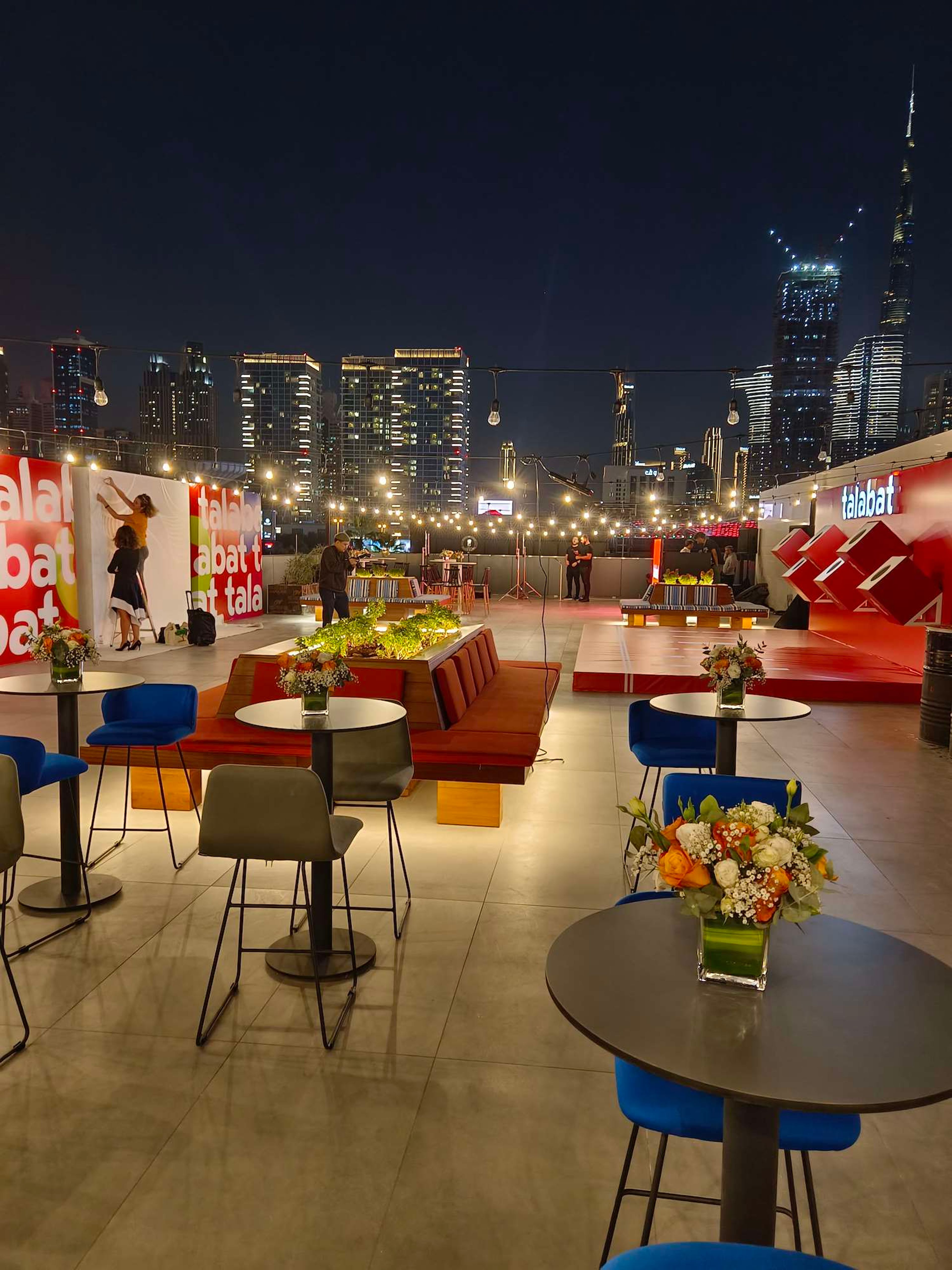 Outdoor night terrace set with lounge chairs, tall tables and flowers for a brand activation, city skyline lit behind