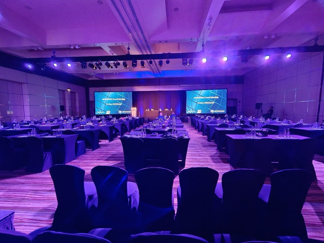 Ballroom with purple lighting and rows of seats facing twin stage screens
