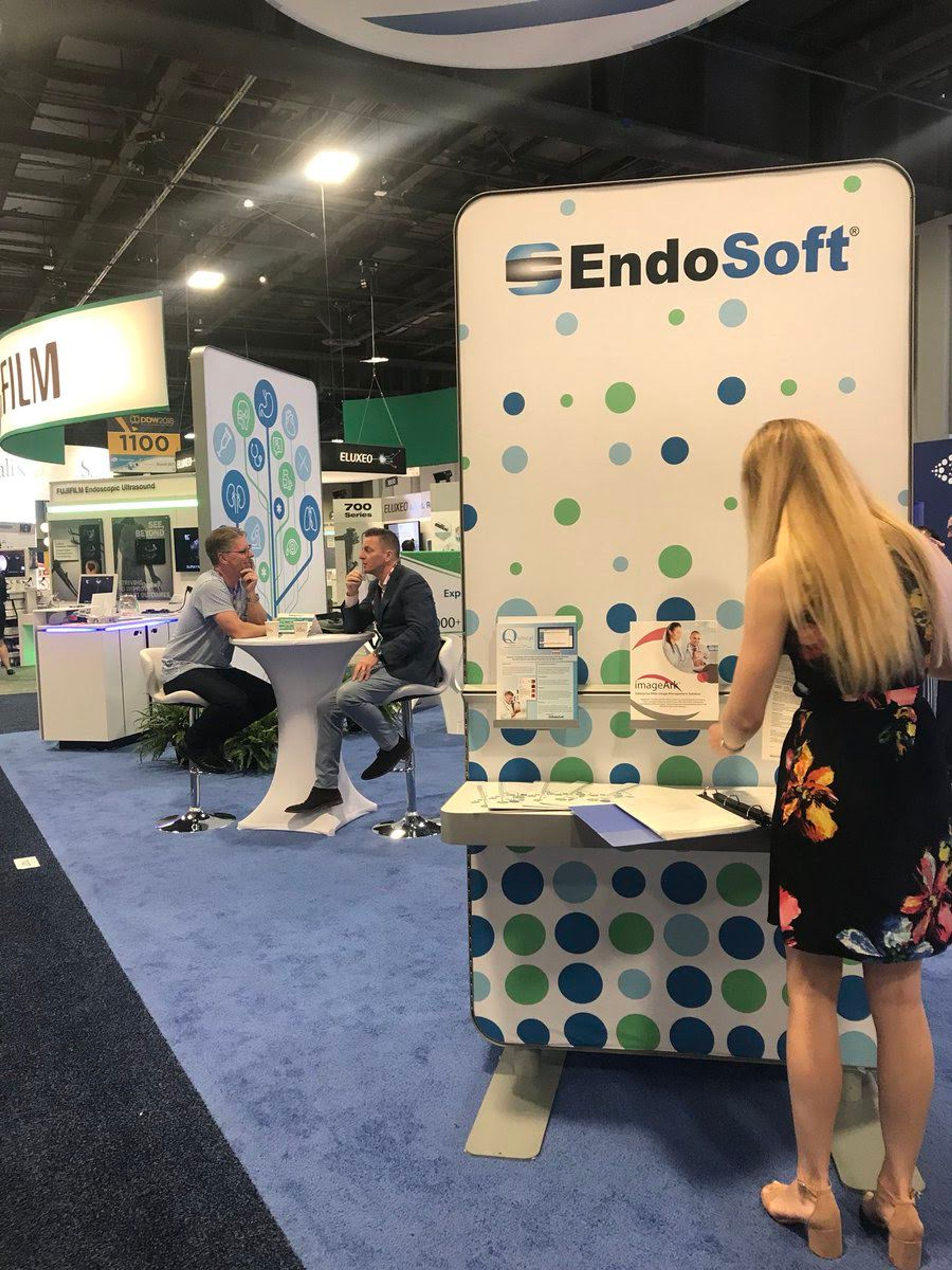 An exhibition booth for EndoSoft, featuring branded signage with a dotted design, while attendees interact nearby. A woman stands reviewing materials, and two individuals are seated at a small table discussing products and services