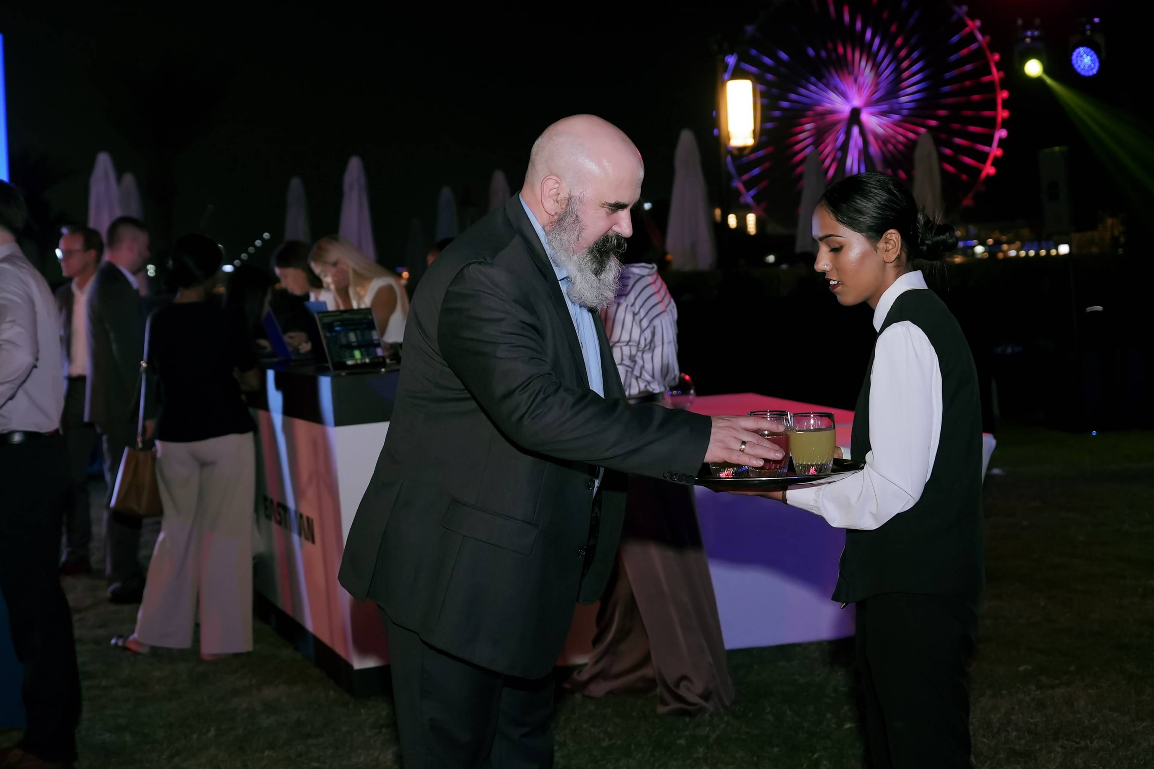 Guest taking a drink from a server at an evening networking reception, with Ain Dubai lit behind