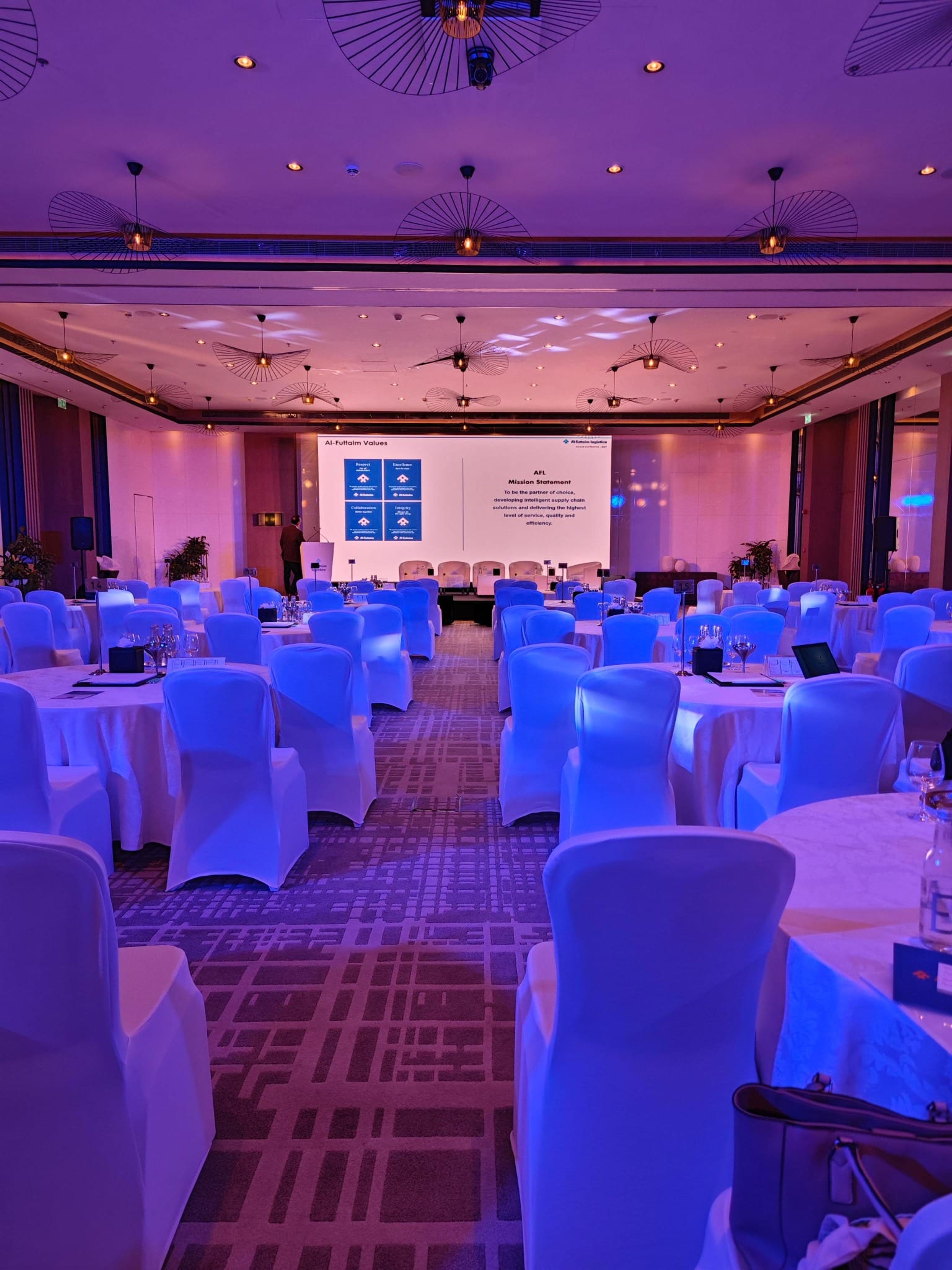 Conference ballroom with white chair covers and round tables facing the stage screen