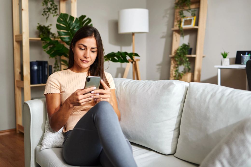 Uma mulher com longos cabelos castanhos está relaxando em um sofá branco e olhando para um smartphone em suas mãos