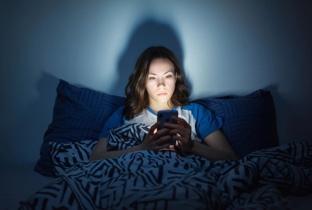Une femme vêtue d'un t-shirt bleu est assise dans son lit et regarde l'écran d'un téléphone portable pendant la nuit.
