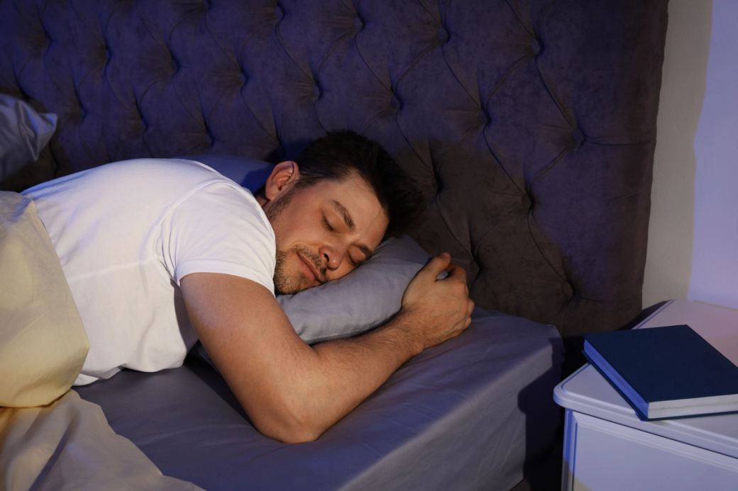 Un homme aux cheveux bruns courts dort profondément dans un lit dont la tête de lit est grise.