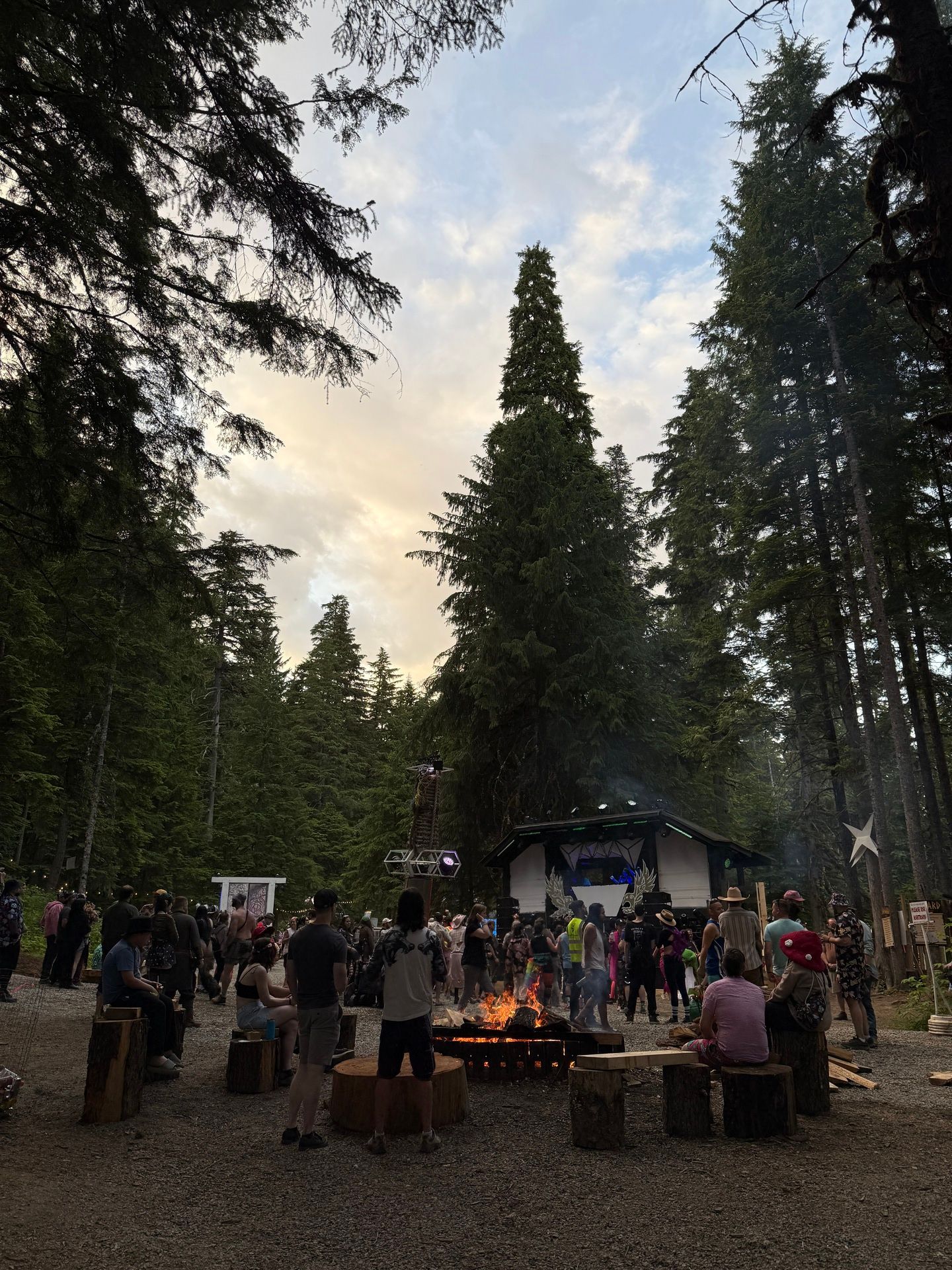 A forest clearing at dusk with festivalgoers gathered around a fire pit, tall evergreens towering overhead and a lit stage behind them.