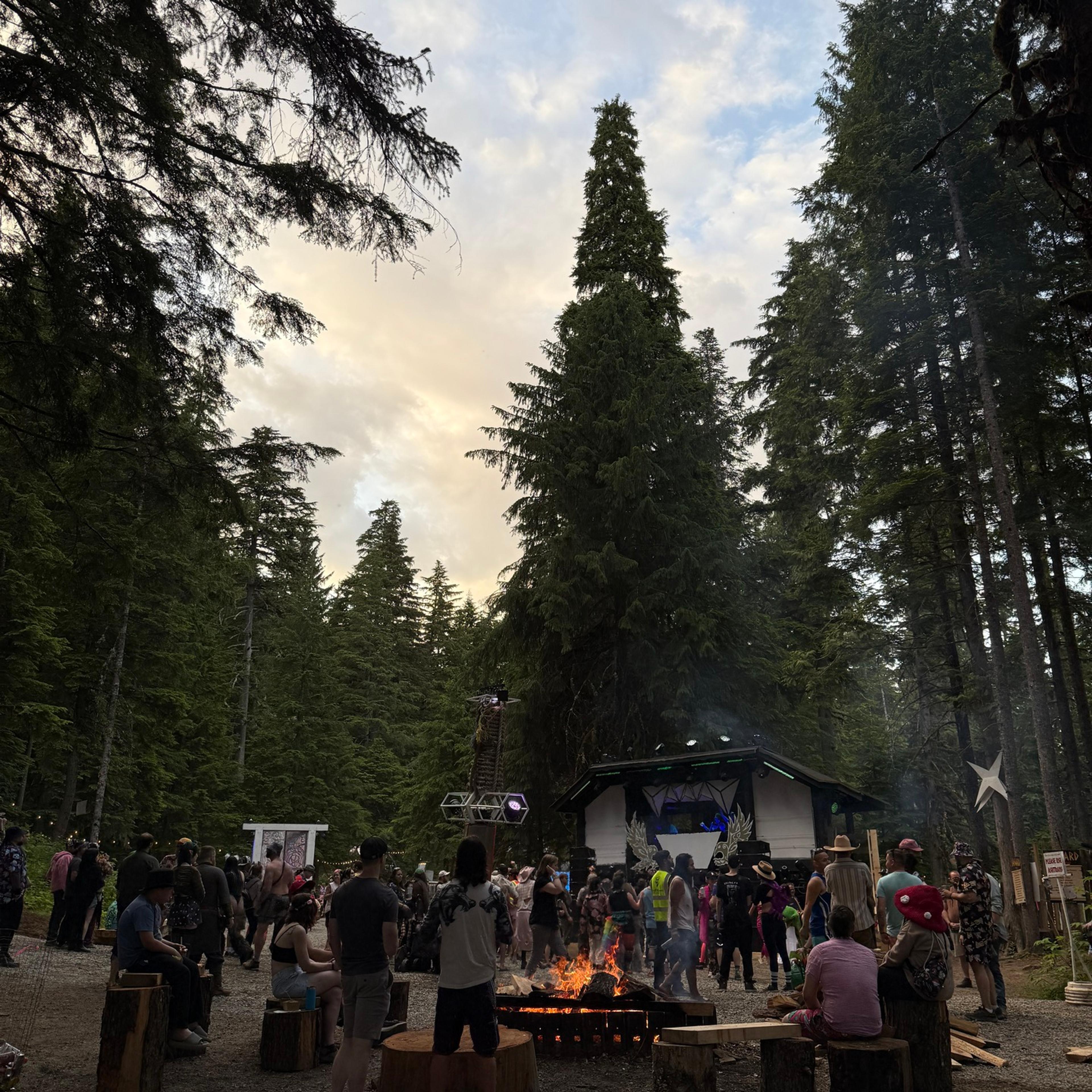 A forest clearing at dusk with festivalgoers gathered around a fire pit, tall evergreens towering overhead and a lit stage behind them.