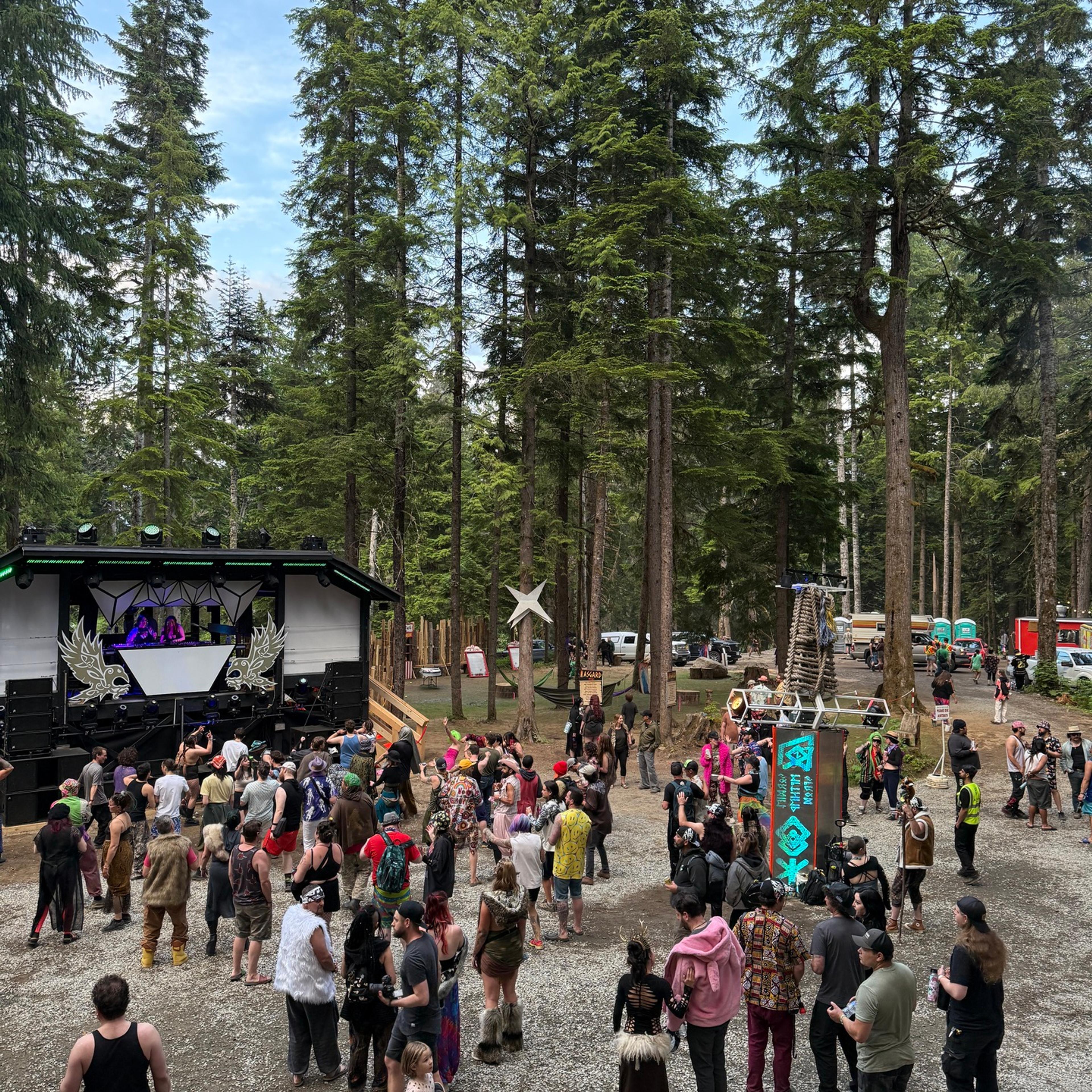 A daytime aerial view of a costumed festival crowd in a forest clearing, with a decorated stage and art installations among the trees.