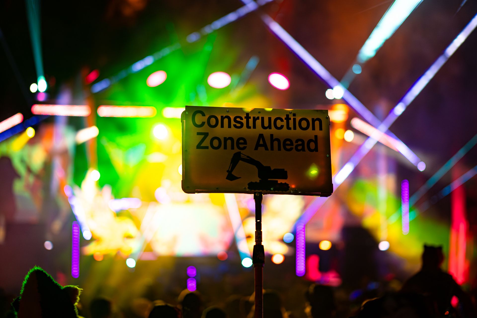 A "Construction Zone Ahead" sign held up in a crowd, colorful stage lights blurred in the background.