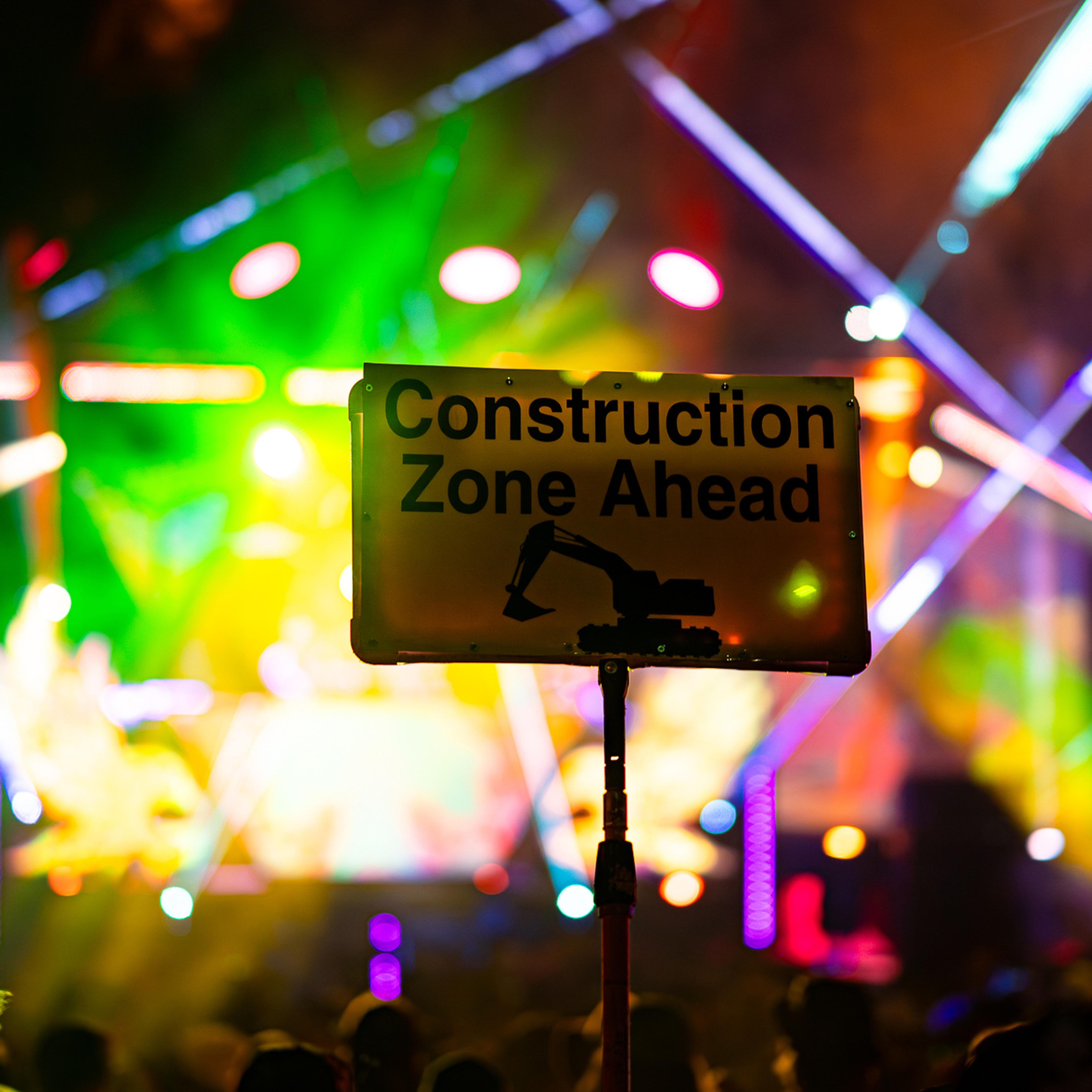 A "Construction Zone Ahead" sign held up in a crowd, colorful stage lights blurred in the background.