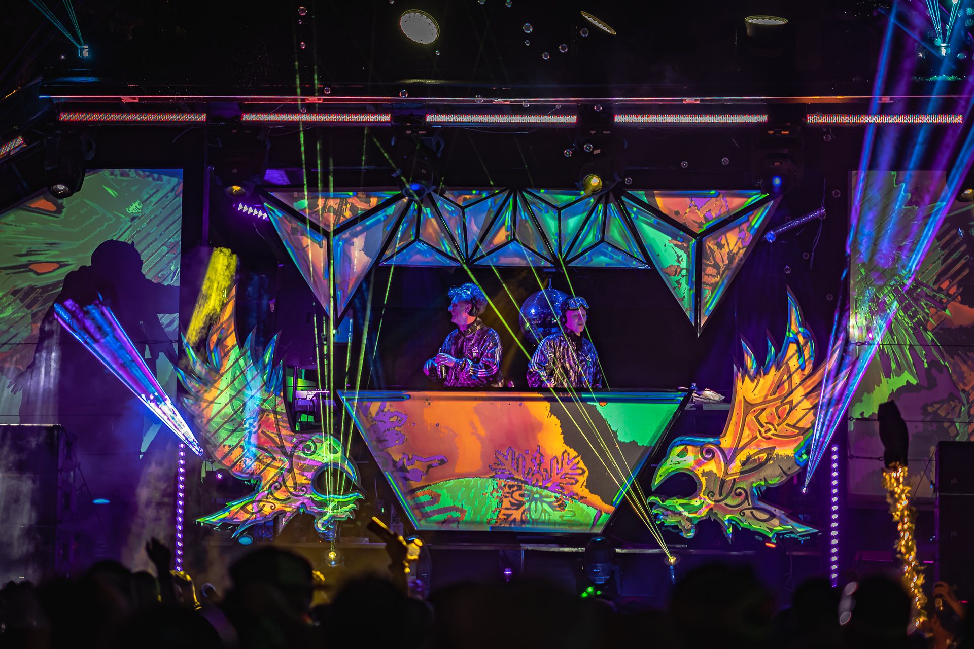 Two performers at a geometric DJ booth surrounded by psychedelic projected visuals and laser beams shooting into the crowd.