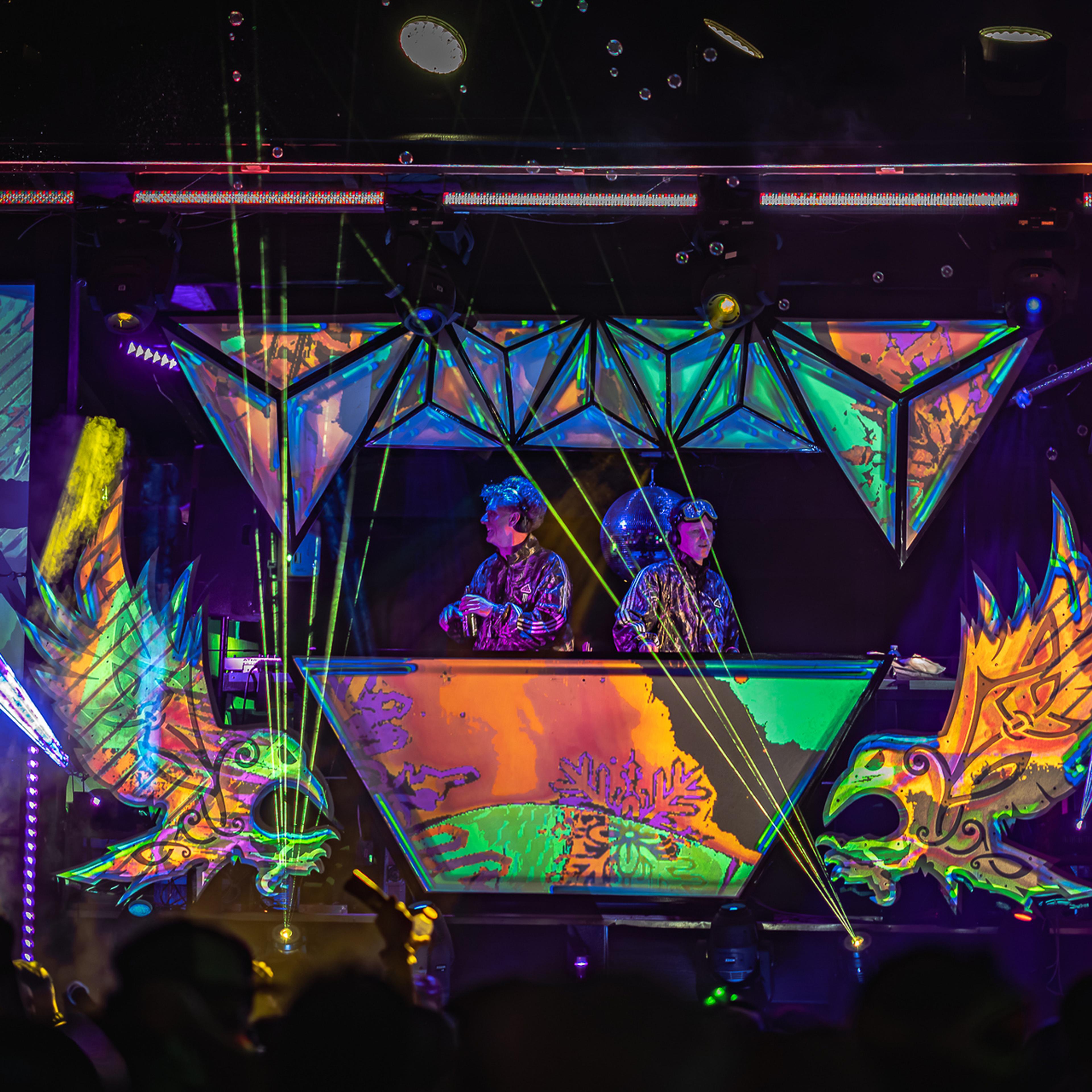 Two performers at a geometric DJ booth surrounded by psychedelic projected visuals and laser beams shooting into the crowd.