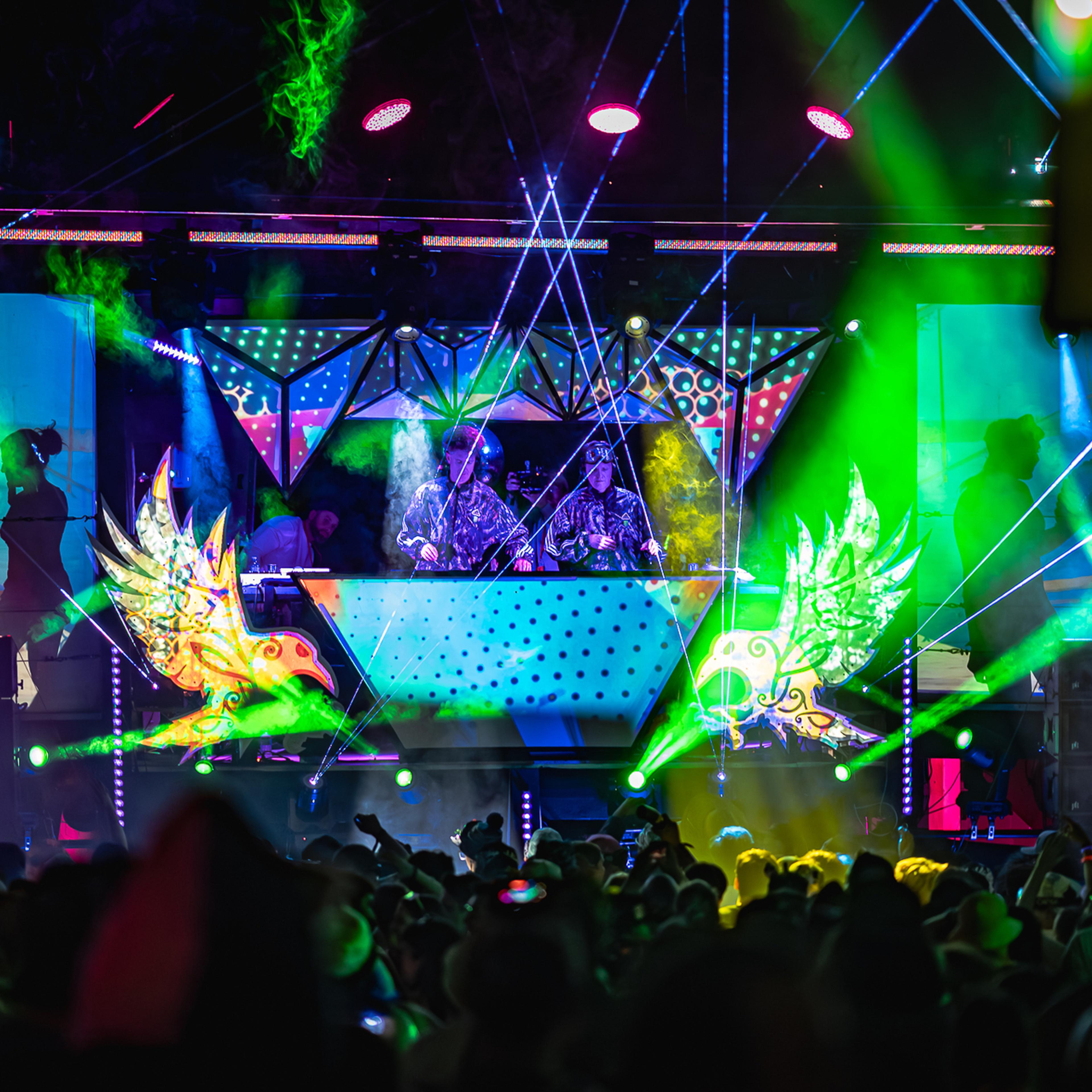 Two DJs performing on a vivid stage with green smoke, laser beams, and colorful winged creature sculptures on either side.