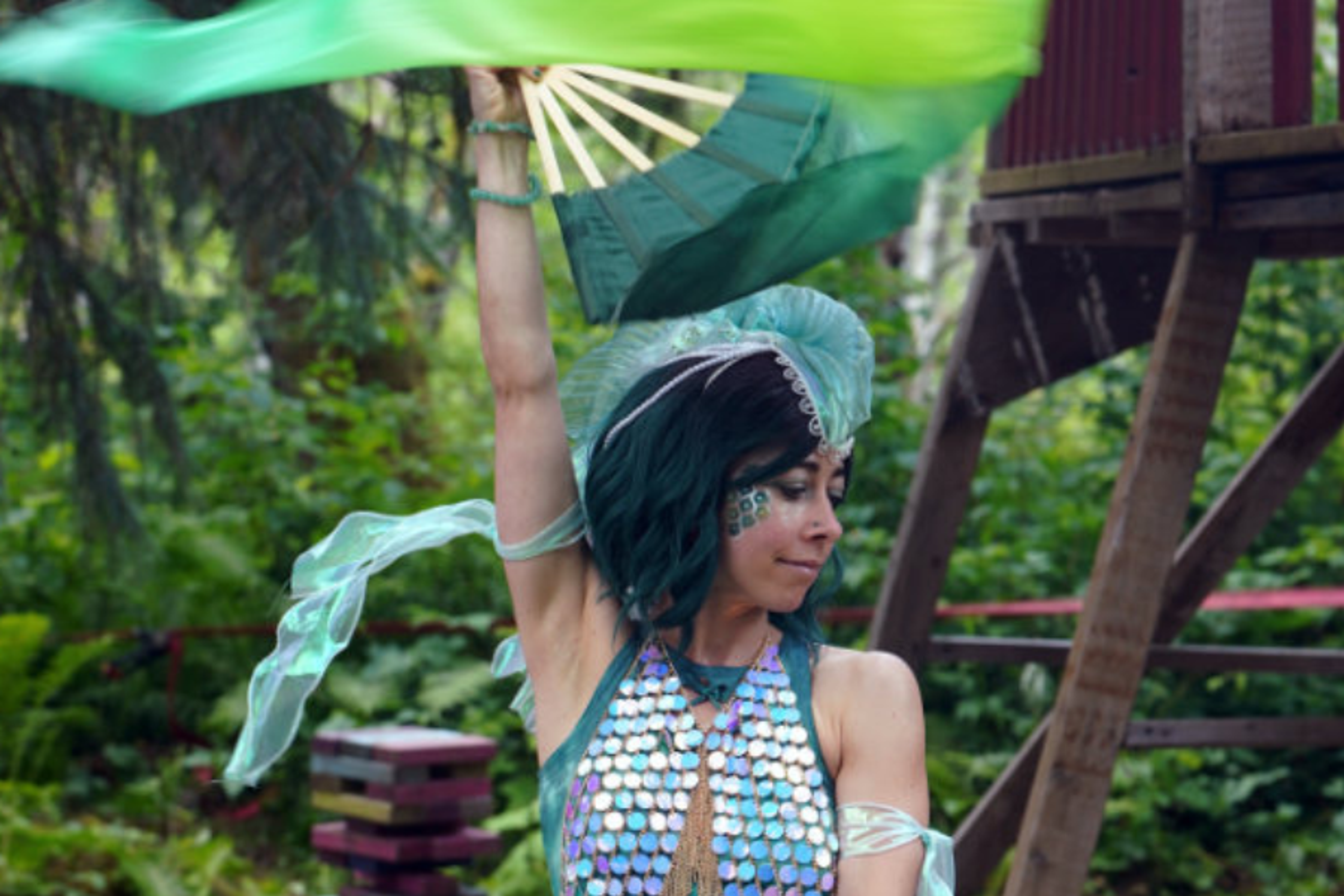 Performer in teal sequined costume and headdress waving silk fans outdoors at ValhallaFest