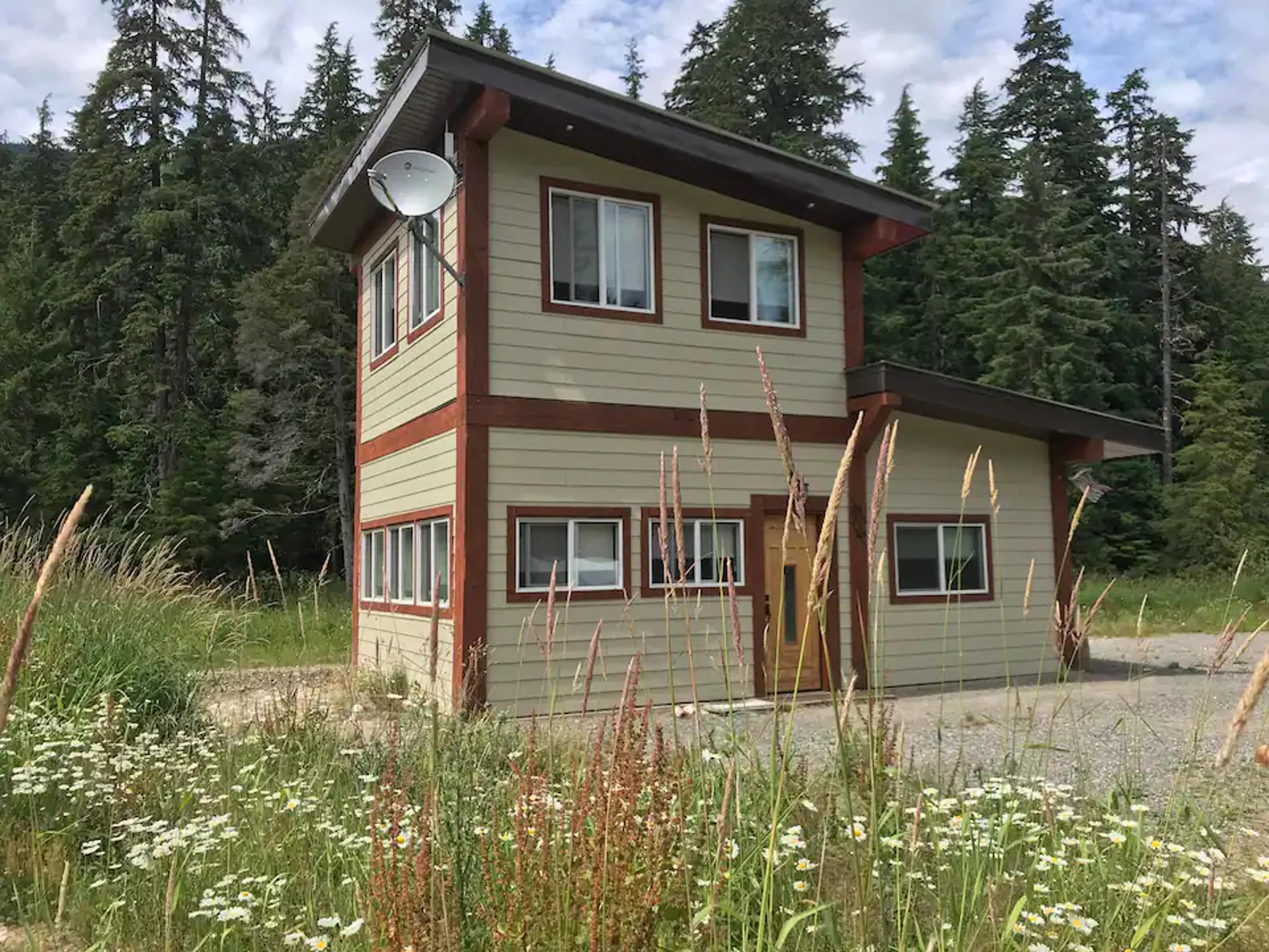  Gemini said  The ValhallaFest Gatehouse, a two-story tan building with red trim and a satellite dish, stands at the edge of a forest clearing surrounded by wildflowers.