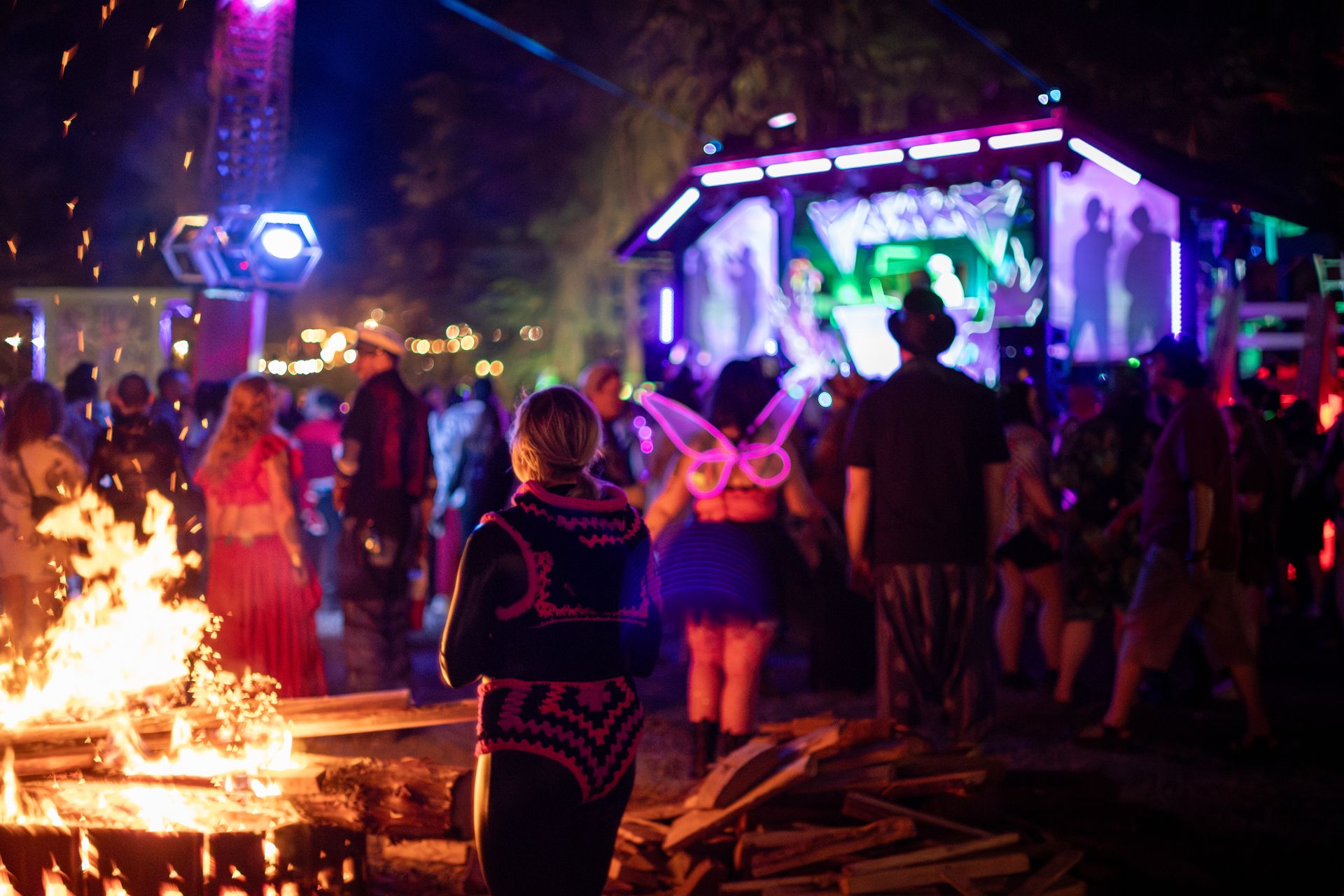 Festival-goers dancing near a bonfire at night, one wearing glowing pink butterfly wings, with a neon-lit stage in the background.