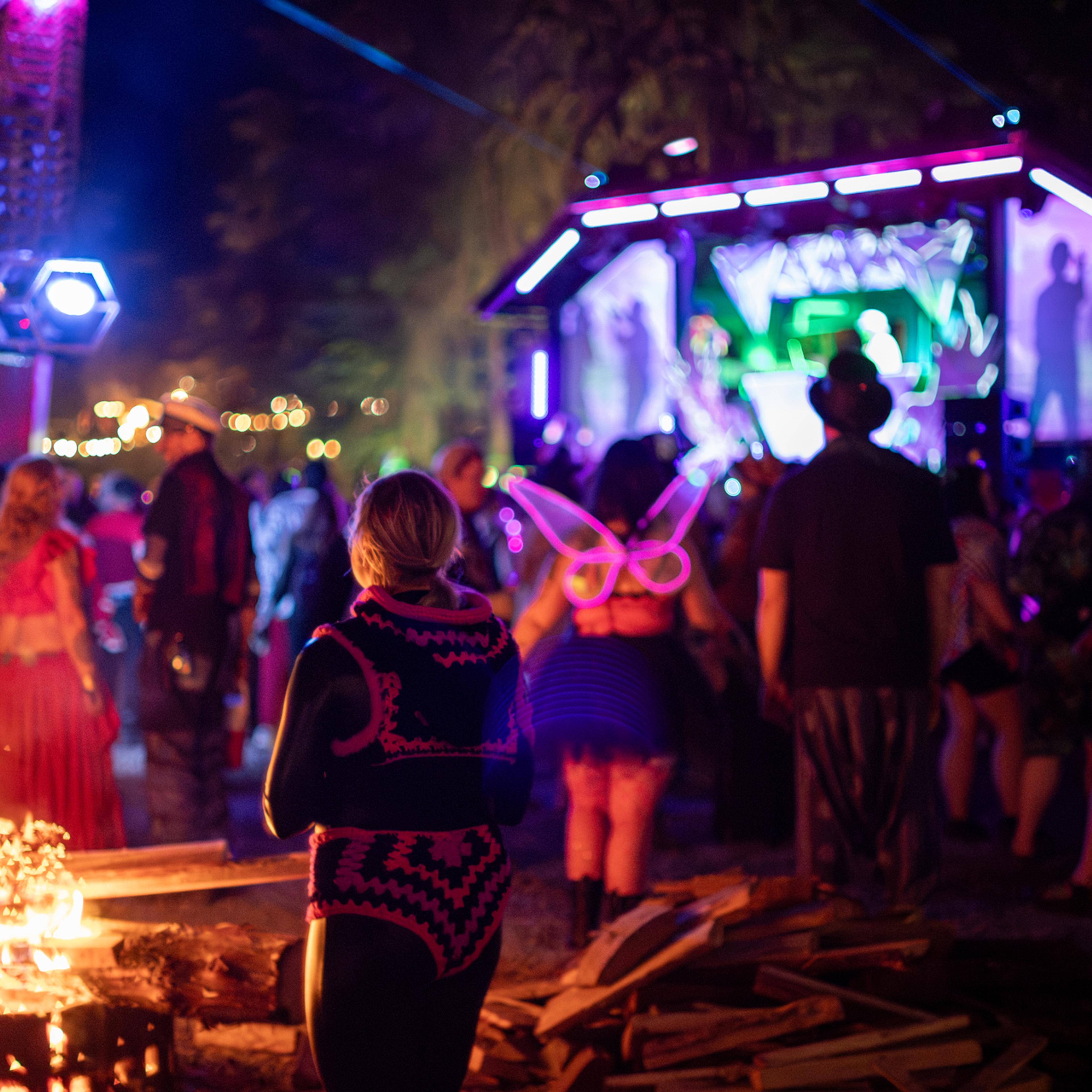 Festival-goers dancing near a bonfire at night, one wearing glowing pink butterfly wings, with a neon-lit stage in the background.
