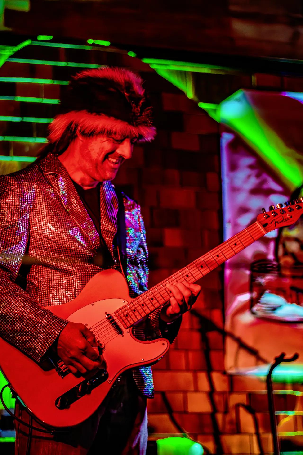 Guitarist in sparkly jacket and feathered hat performing under colorful lights on AgeHa stage.