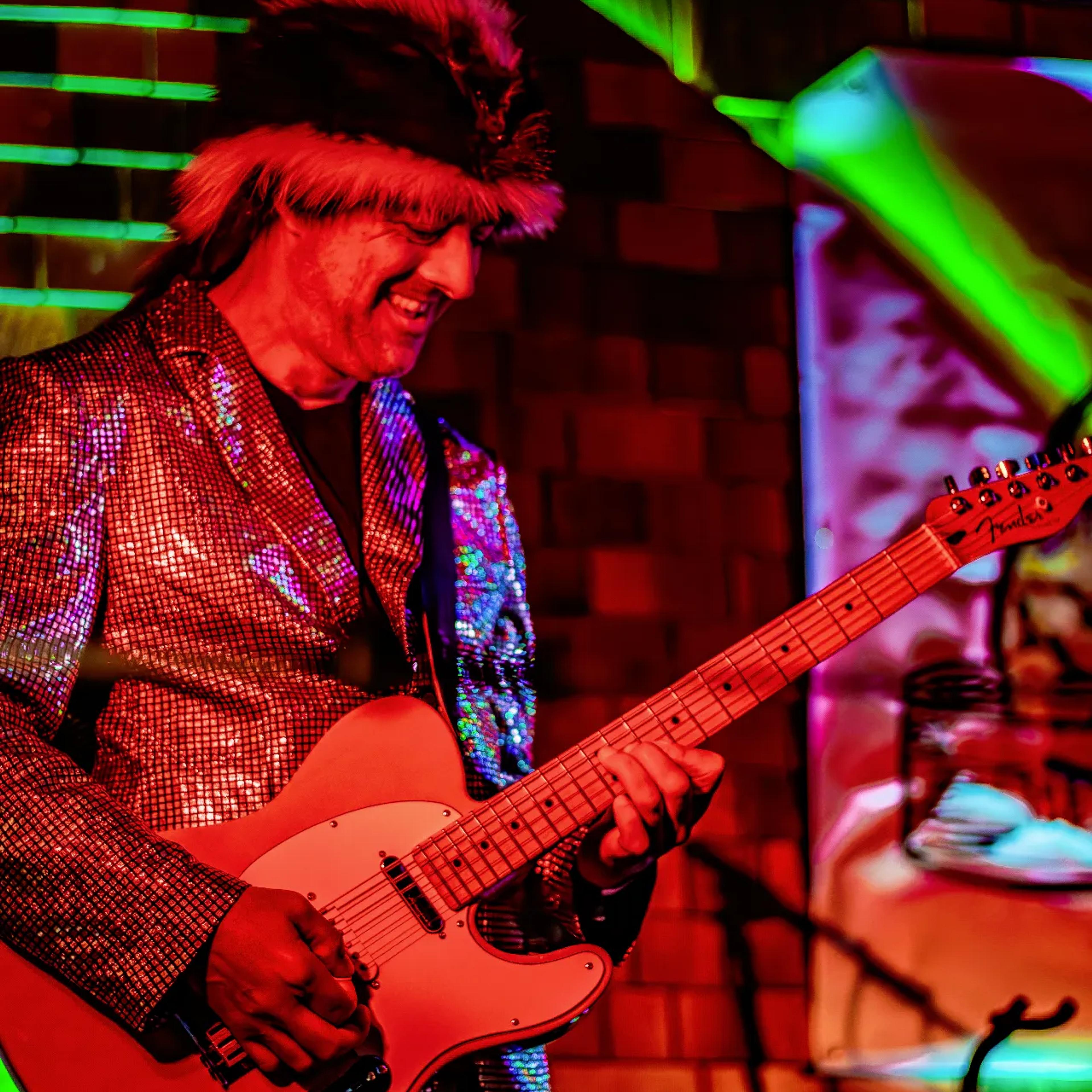 Guitarist in sparkly jacket and feathered hat performing under colorful lights on AgeHa stage.