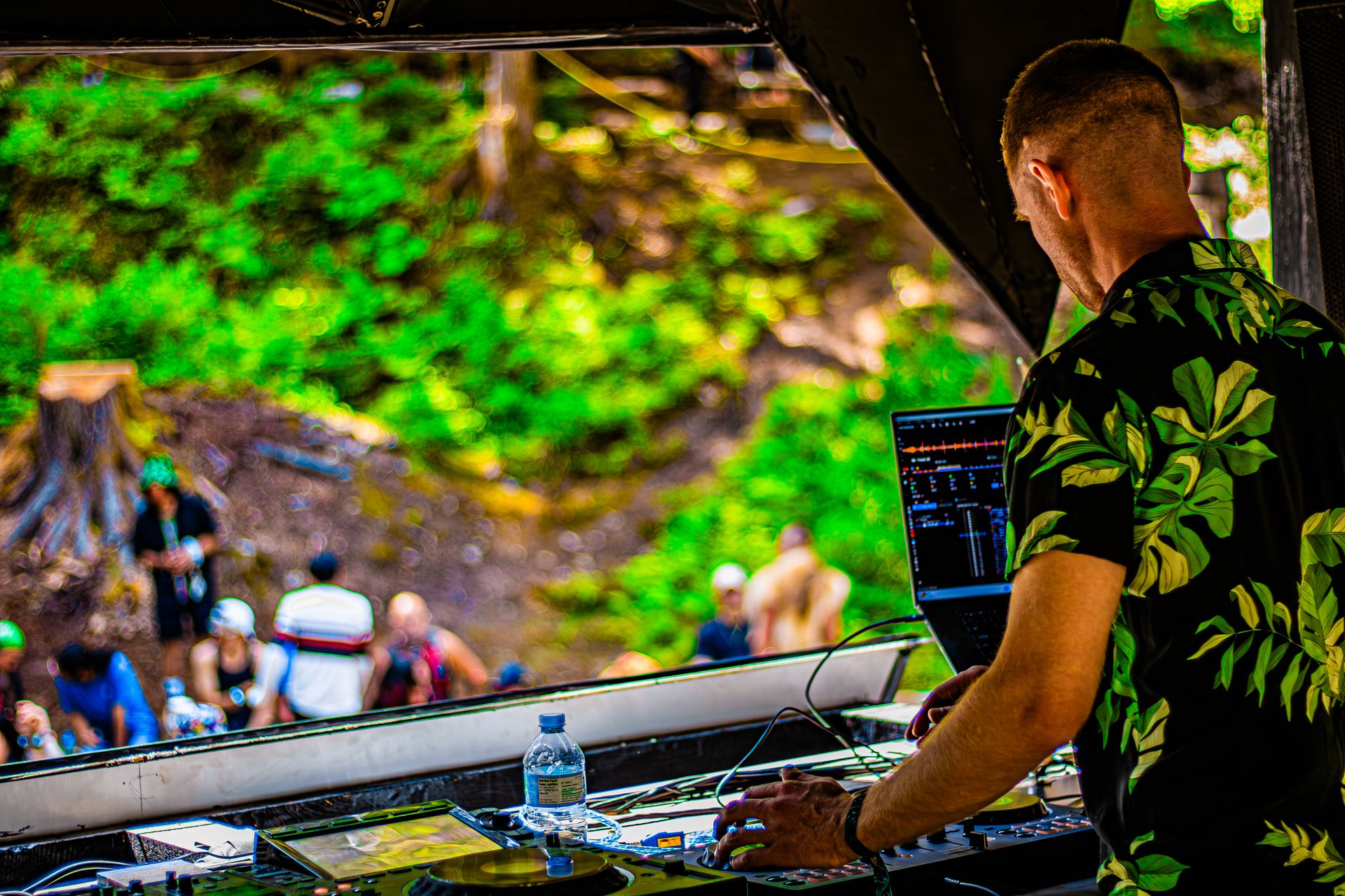 A DJ in a floral shirt works the decks outdoors, lush green forest and seated crowd blurred behind him.